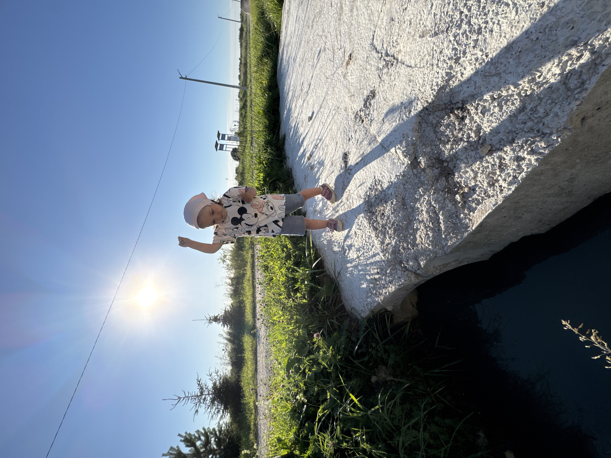 In Kempelevo, Russia, a young child wearing a pink bandana, a t-shirt with Mickey Mouse designs, and grey shorts stands on a concrete structure. The child is raising one arm, appearing to wave or gesture. The concrete structure extends over dark water, with green grass and vegetation on the left side of the frame. In the background, a bright sun shines in a clear blue sky, casting long shadows on the concrete. A dirt path is visible behind the grass, with trees and utility poles further in the distance. The scene appears to be outdoors during daylight hours.