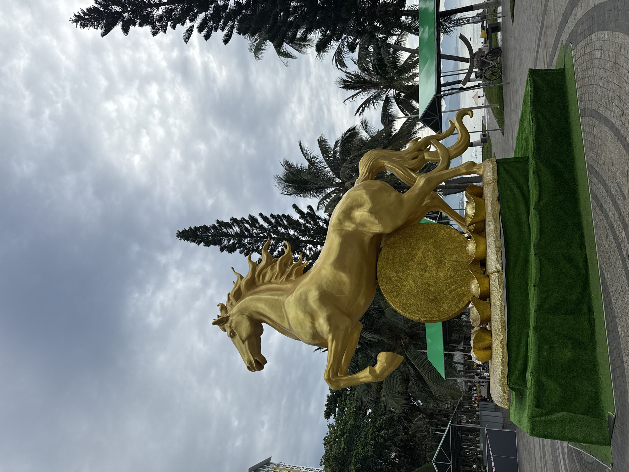 The image shows a golden horse statue in a city park in Nha Trang, Vietnam. The statue is depicted in a dynamic pose, with its front legs raised and its mane flowing. It stands on a large, circular golden disc, which is in turn supported by a tiered base covered in artificial grass. In front of the golden disc, several golden ingots are arranged.
In the background, palm trees and tall pine trees are visible, suggesting a tropical or subtropical climate. Beyond the trees, a body of water, likely the sea, can be seen, with distant hills or mountains on the horizon. There are also some small structures with green roofs and a building in the far left, indicating an urban setting. A bicycle is parked near a bench in the middle ground, and a few people are visible in the distance near the water. The sky is overcast with clouds. The overall scene appears to be a public space, possibly a promenade or a plaza, decorated for a festive occasion, given the prominent golden decorations.