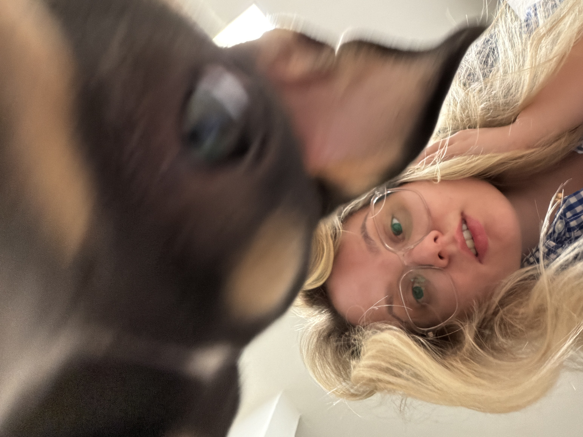 A low-angle shot captures a woman and a dog. The woman, positioned towards the right, has blonde hair, fair skin, and wears clear-framed glasses. Her expression is neutral, and she is wearing a blue and white patterned top. The dog, dark in color, occupies the left side of the frame, its face blurred and close to the camera. The background is indistinct but appears to be an indoor space with white walls. There is no evidence that the picture was taken in Tel-Aviv, Israel.