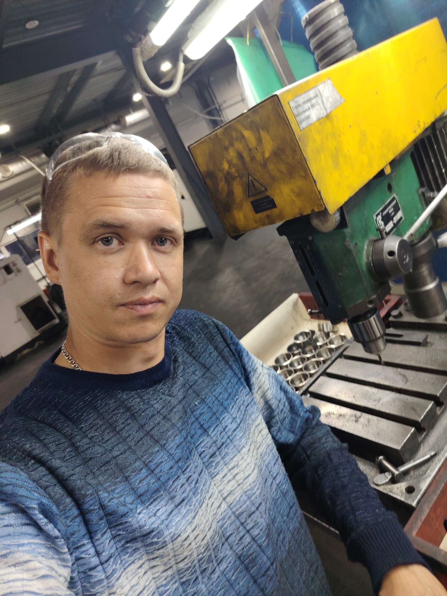 A male individual, appearing to be in his late 20s to early 40s, is captured in a selfie within an industrial workshop. He has short, light-colored hair and is wearing clear safety glasses perched on his head. His eyes are blue, and he maintains a neutral expression while looking directly at the camera. He wears a blue and white patterned long-sleeved sweater with a textured, diamond-like design, and a silver chain is visible around his neck.
To the right of the man is a prominent yellow and green industrial drilling machine. A label on the yellow housing, written in Russian, identifies it as "Станок сверлильный настольный ДМ12" (Desktop Drilling Machine DM12) with inventory number "Инв. №005907". Another warning label with a triangular hazard symbol reads "Осторожно! Движущиеся части!" (Caution! Moving parts!). The machine features a visible drill chuck and bit, positioned above a metal work table with grooves. Adjacent to the work table, a white tray holds multiple small, circular metallic components.
The background reveals a typical industrial environment with visible ceiling structures, bright overhead fluorescent lighting, and other manufacturing equipment, including a partially visible white machine on the left. The scene is consistent with a factory or workshop in Russia, in the city of Noviy.