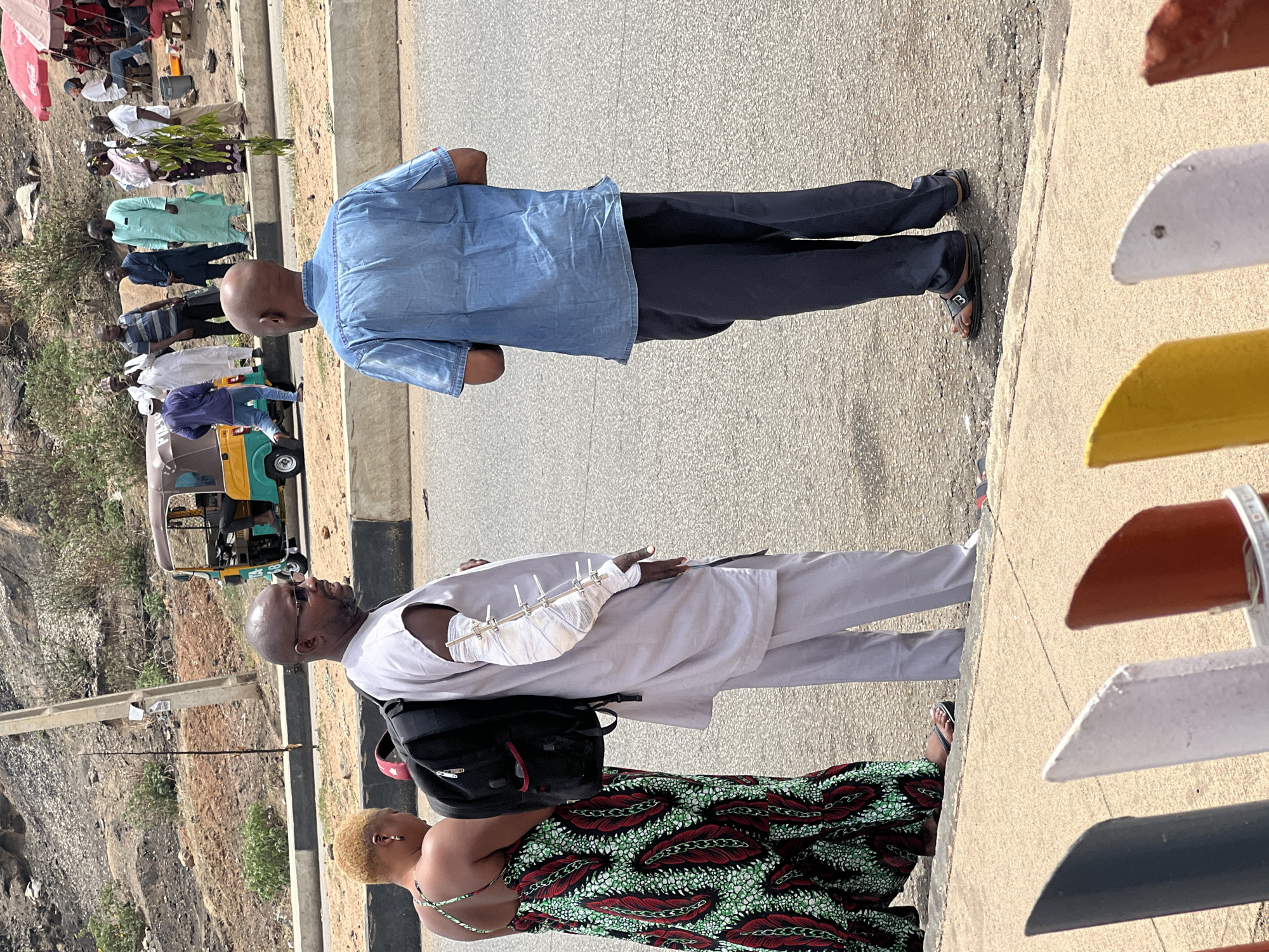 The image, oriented correctly, captures an outdoor scene featuring several individuals, likely in Jos, Nigeria. In the immediate foreground, a bald man wearing a light blue denim shirt and dark trousers stands with his back to the camera. Beside him, another bald man, wearing sunglasses and a white traditional outfit, is prominent due to a significant injury to his left arm, which is heavily bandaged and supported by an external fixator with visible metal rods. He also wears a black backpack. To his left and slightly behind him is a woman with short, blonde hair, dressed in a vibrant, patterned African fabric dress (Ankara) featuring deep green, red, and white motifs.
In the mid-ground and background, numerous other people are visible, some appearing to be queuing or gathered along a dirt path or roadside. Many are dressed in traditional Nigerian clothing. A green and yellow Keke (auto-rickshaw) is parked in the mid-ground. The overall setting includes a paved road or path in the foreground, a concrete curb, and an arid, rocky landscape with sparse vegetation in the distant background, typical of some regions in Nigeria.
The activity appears to be a gathering or waiting, possibly at a public transport stop or a local market area. The man with the prominent arm injury suggests a recent incident or an ongoing medical situation.
The scene is illuminated by bright daylight, casting clear shadows, indicating midday or early afternoon. Visible text includes "BILAL" and "21" on the Keke, and a "Coca-Cola" logo on a red umbrella in the far background.
