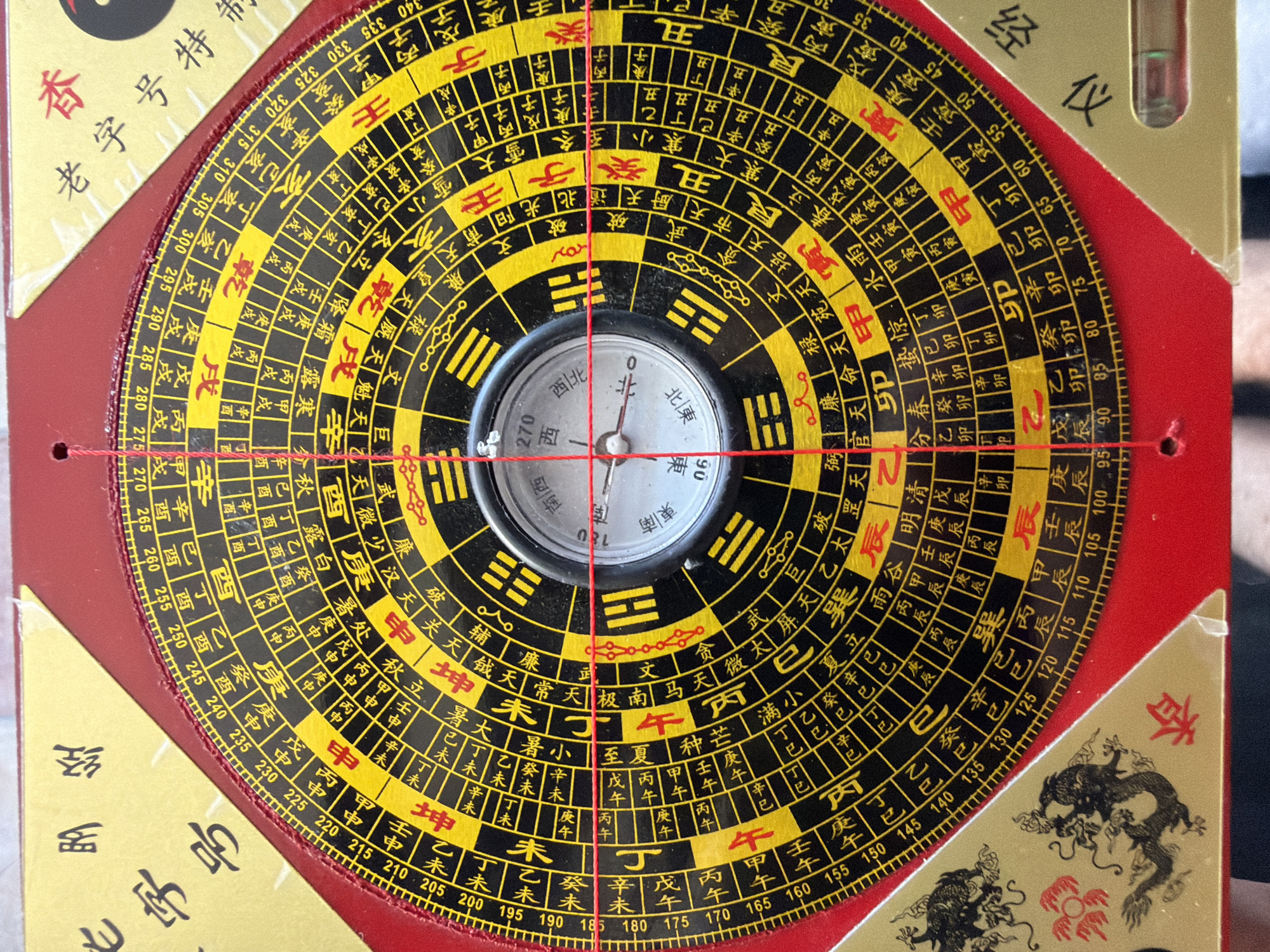 The image shows a Chinese Feng Shui compass, also known as a Luopan. It features concentric rings filled with Chinese characters and symbols, surrounding a central magnetic compass for orientation. The compass is red and gold in color. The needle of the compass is aligned, pointing at north. It's surrounded by a level. The compass is overlaid by red string aligned on the North/South and East/West axes. Some parts are visible on the left, right and upper side showing a square box with what appears to be chinese characters in red. A portion of a box featuring a dragon is visible at the lower right corner. The image does not show Budva, Montenegro or contain any indicators of that location.