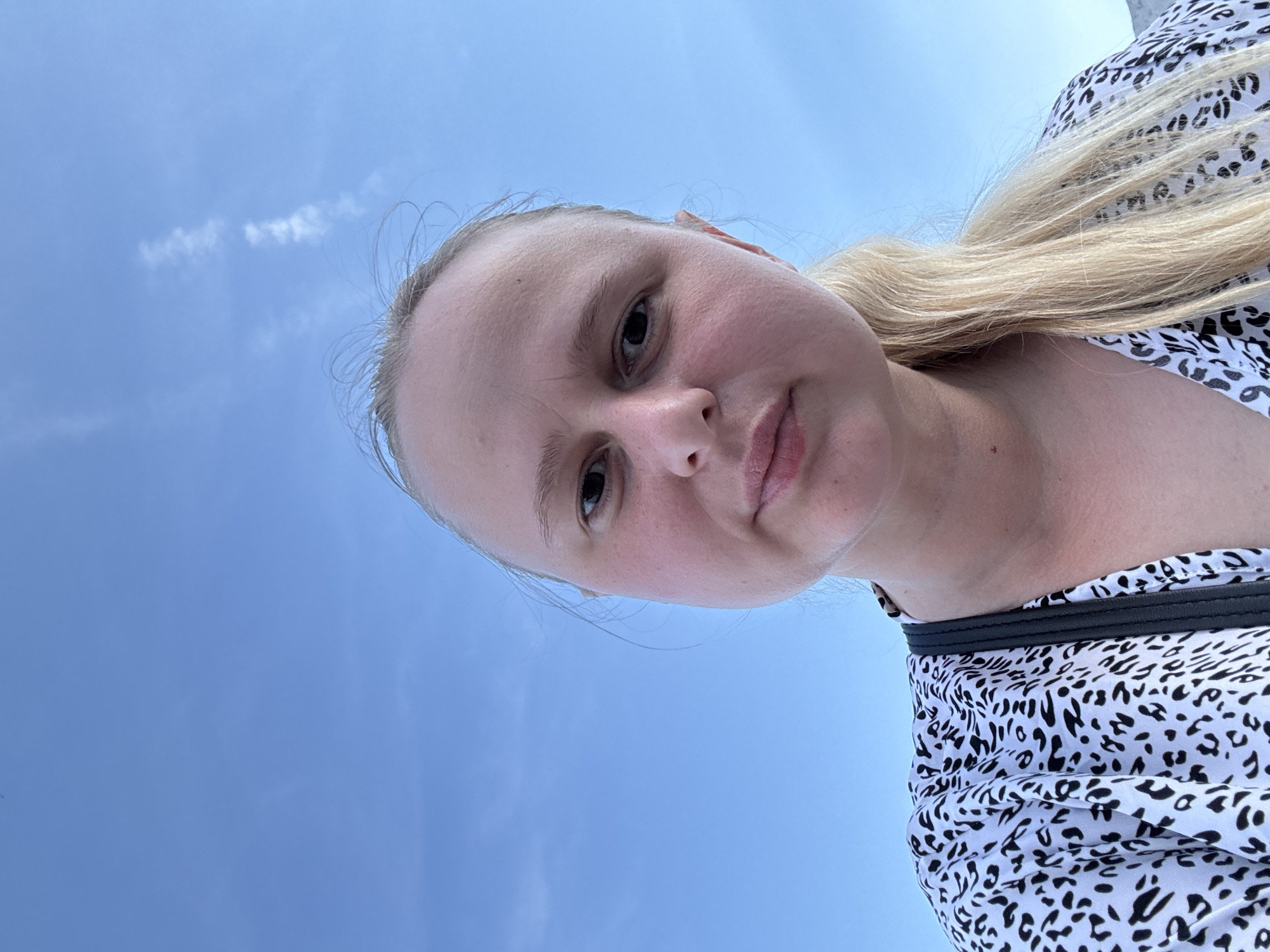 A close-up, eye-level, full shot shows a young Caucasian woman with blonde hair pulled back into a ponytail, facing the camera. She is wearing a black and white leopard print top. Her skin is fair with some redness on her cheeks. The background is a clear blue sky with a few wispy white clouds. The image appears to have been taken outdoors during the day. There is no discernible indication of the location being Saratov, Russia.