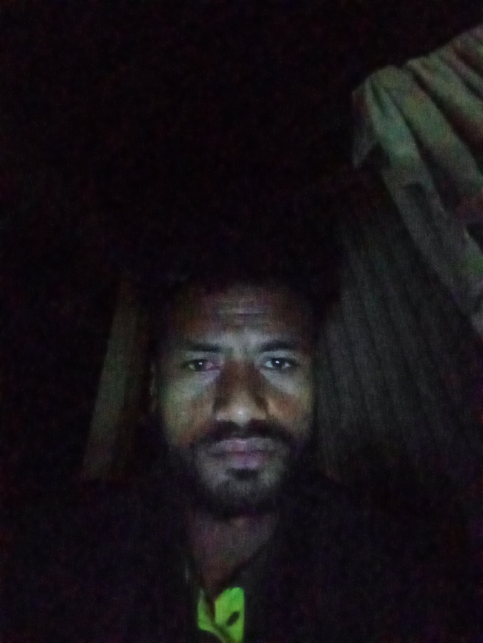 A man with dark skin and a beard is in the center, dimly lit. He's wearing a dark shirt with a neon green design visible at the collar. The background is dark, but some indistinct shapes suggest either curtains or a draped fabric to his right and a wooden board to his left. The image quality is grainy.