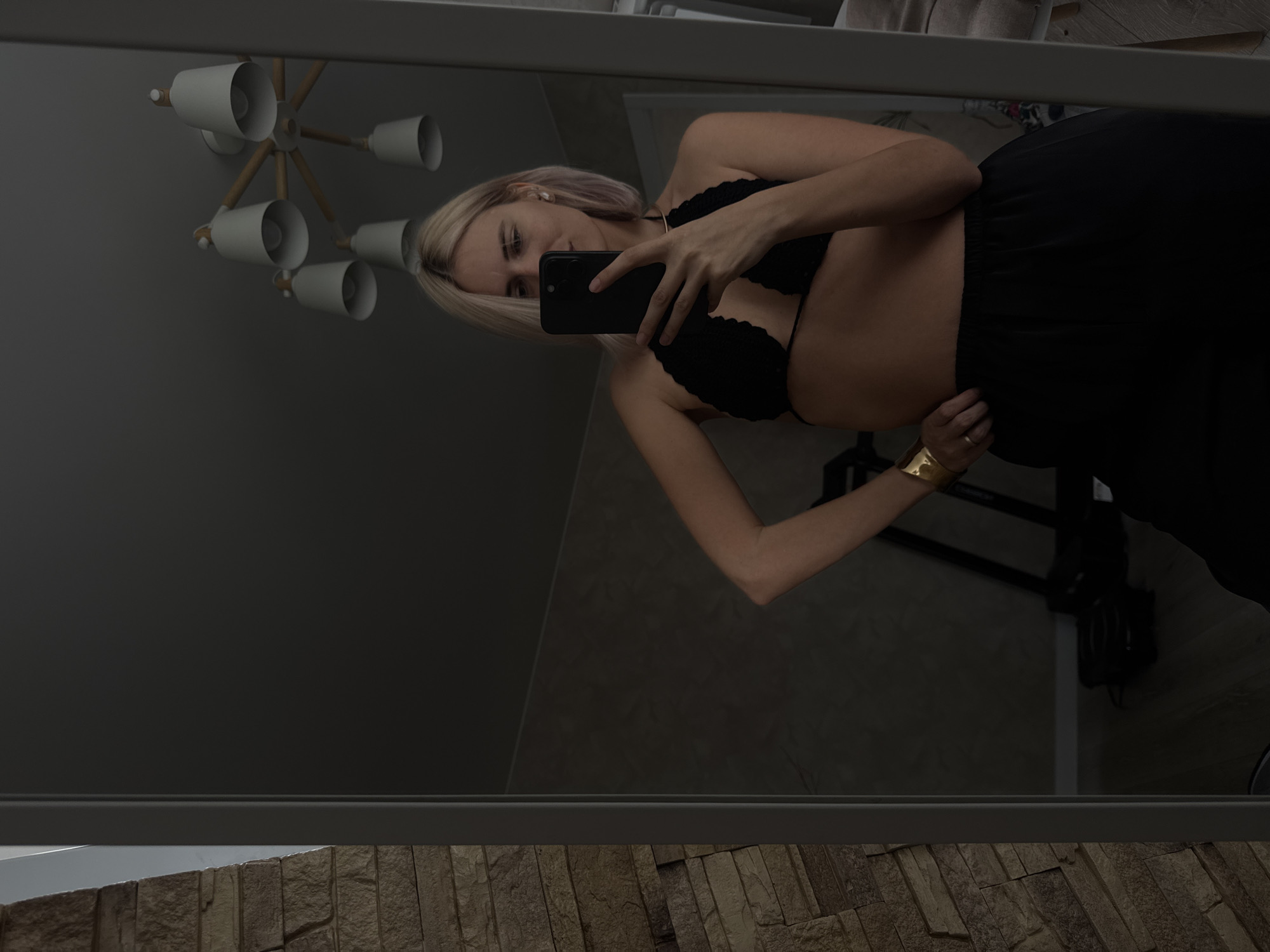 A fair-skinned woman with blonde hair is taking a selfie in a mirror. She is wearing a black bralette and loose black pants. She is holding a black smartphone and her left hand is resting on her hip, where a wide gold cuff bracelet is visible. The reflection in the mirror shows a modern chandelier with white shades and wooden arms, a gray wall, and what appears to be a stone wall at the bottom of the reflection. The location is not discernible from the image, and the city of Saratov, Russia, cannot be confirmed.