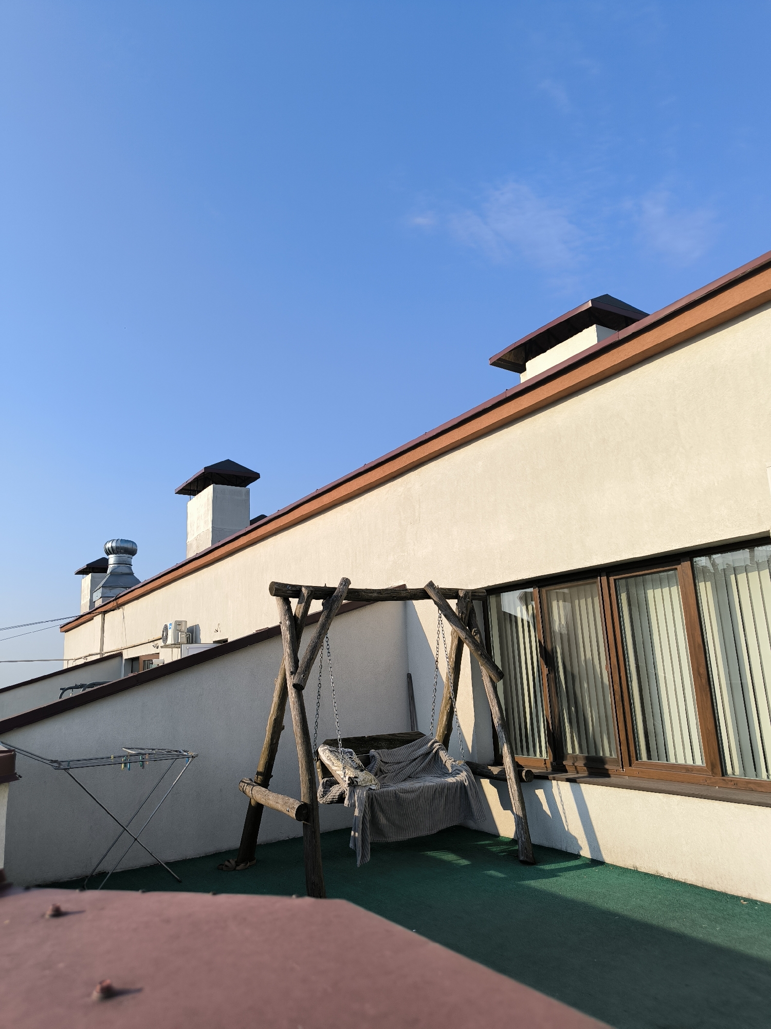This is an outdoor scene on a rooftop in Chișinău, Moldova, under a clear blue sky. The foreground features a portion of a reddish-brown structure. Dominating the mid-ground is a wooden swing set with a cushioned seat covered in a grey textile. To the left of the swing is a foldable clothes drying rack with several blue clothespins. The background shows a white building with a brown-trimmed roofline and several chimneys or ventilation pipes protruding from it. A large window with wooden frames and vertical blinds is visible on the building's facade. The rooftop area is covered with a green carpet-like surface. The sun is casting shadows, indicating it is likely late afternoon. There are no people or animals present in the image.