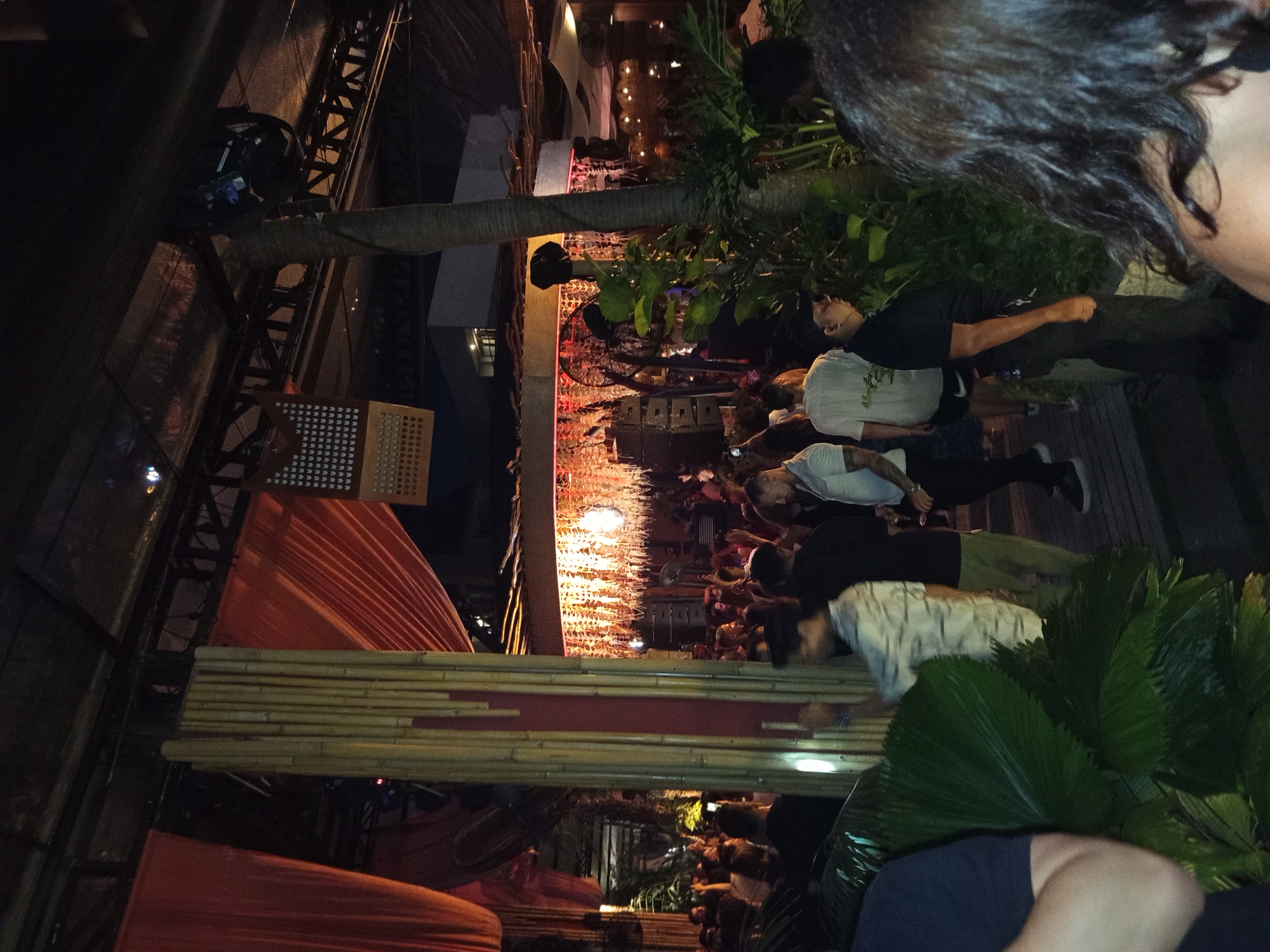 This image captures a vibrant, dimly lit outdoor nightlife venue in North Kuta, Indonesia. The scene is populated by a dense crowd of people engaged in social interaction. Overhead, a robust structural canopy features dark metal trussing, from which large panels of draped orange fabric are suspended. Decorative elements include a vertical column clad in bamboo poles with a red stripe, and a perforated gold-colored panel adorned with multiple heart-shaped cutouts.
In the mid-ground, numerous individuals, casually dressed in t-shirts, polo shirts, and shorts, are standing and mingling. Several large tropical plants, including broad fan palms and other green foliage, are strategically placed throughout the venue, partially obscuring views and contributing to a tropical aesthetic. A prominent palm tree trunk stands to the right of the center.
Towards the background-center, a raised area or stage is visible, featuring tall stacks of audio speakers and an elaborate ceiling-like installation of dried, light-colored fronds or leaves, illuminated by numerous small warm lights. This area appears to be a focal point for the crowd. A security guard in a black polo shirt and dark pants, equipped with keys, is visible standing towards the bottom right. The ground surface appears to be constructed of wooden planks. The overall ambiance suggests a lively evening event or gathering.