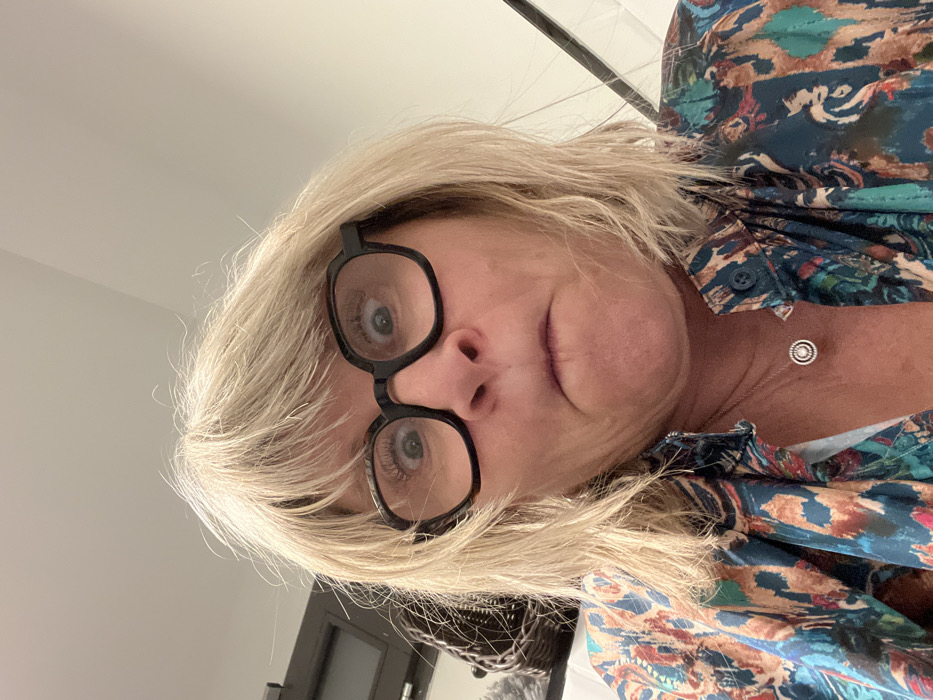 A mature woman with short blonde hair and black-framed glasses looks directly at the camera. She wears a patterned button-down shirt in shades of blue, green, and orange. A simple silver necklace with a circular pendant adorns her neck. The background includes a white ceiling and a glimpse of a dark door and woven basket. The image is rotated.