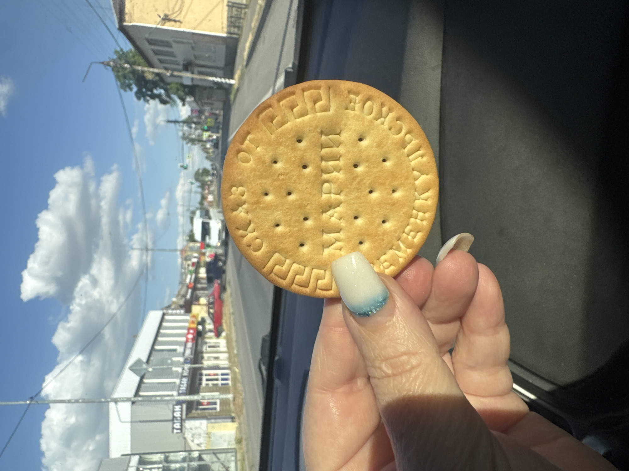 A medium shot captures a person's hand holding a round biscuit with embossed text, positioned in the foreground against a blurred background of a street scene. The biscuit, golden brown and slightly textured with small perforations, displays Cyrillic lettering. The text appears to read "МАРИИ" in the center, and "ОТЕЧЕСТВЕННОЕ" and "ВЕЧНОЕ" around the perimeter. The person's hand is fair-skinned with a manicured nail that features a white base coat with a blue glitter gradient near the tip. The background reveals a street lined with buildings, some with storefronts, and a few vehicles, including a white van and a red car. A traffic light is visible in the distance, suggesting an urban or town setting. The sky is blue with scattered white clouds. The provided location information specifies Kagal'nitskaya, Russia.