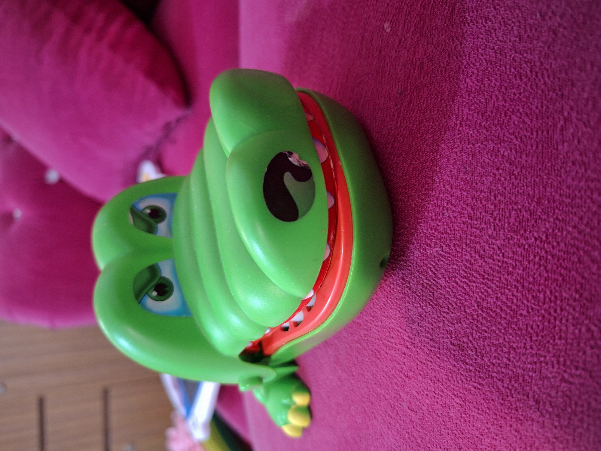 The image presents a vibrant green plastic toy, resembling a cartoonish crocodile or alligator, resting on a bright magenta-colored, textured fabric surface. The toy is depicted with its head and widely-opened mouth facing the right side of the frame. Its mouth features a red inner lining and small, pointed white teeth. Two large, cartoonish eyes are visible; one has a clear black pupil with a white highlight and a blue iris area, while the other is partially obscured but shows a distinct black, crescent-shaped marking. Small, yellow-tipped feet are visible at the bottom of the toy.
The background primarily consists of the magenta fabric, which shows a prominent woven texture. To the left, a blurred magenta object, possibly a tufted cushion or part of a sofa, adds depth. A small portion of a wooden surface is visible at the very bottom left. The scene is stationary, with no specific activity taking place. The colors are bright and saturated, with soft, even lighting suggesting an indoor setting. There is no visible text in the image. The setting is indoors in Ciseeng, Indonesia.