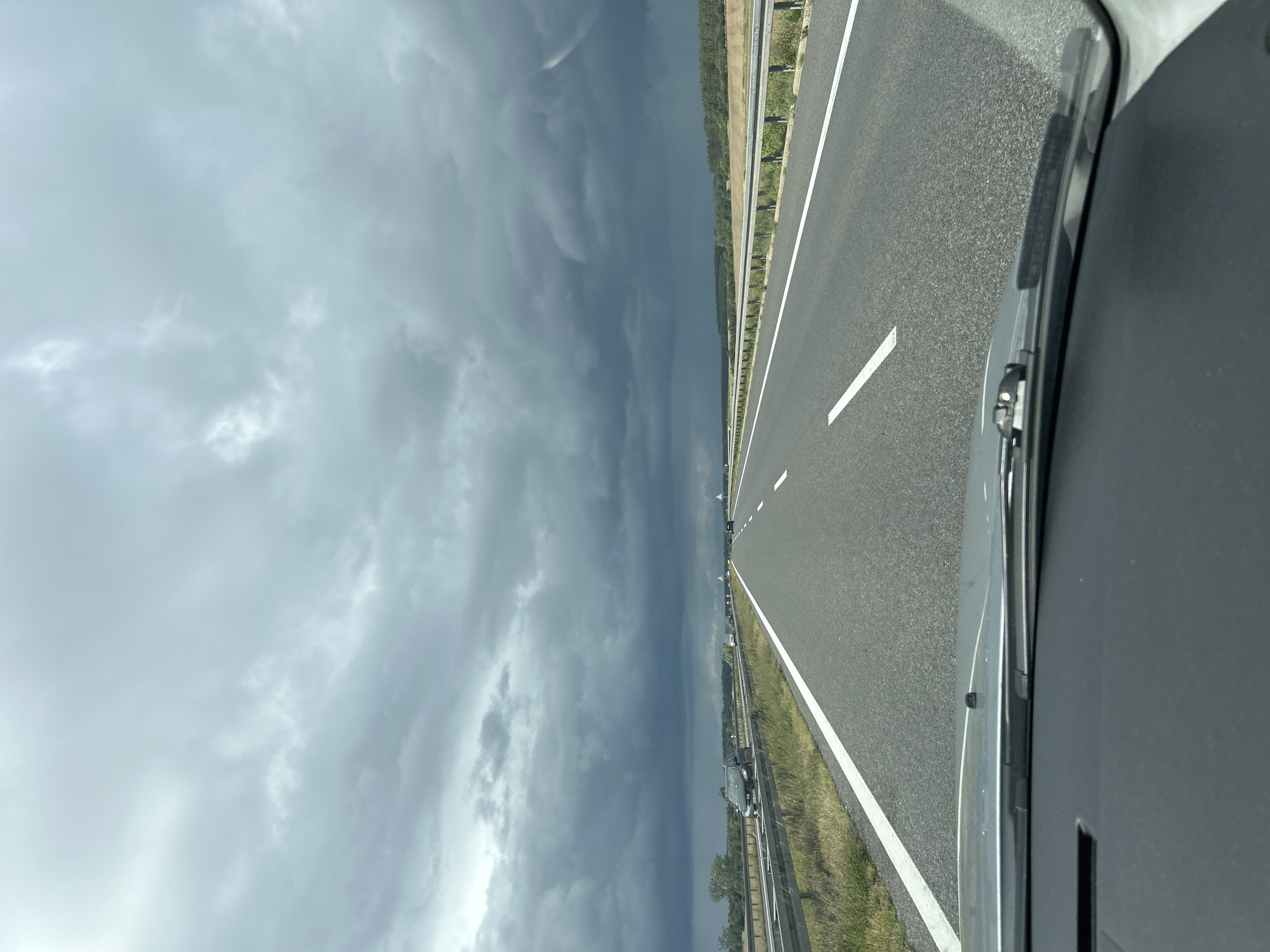 The image shows a view from inside a vehicle traveling on a highway. The road is asphalt with white lane markings. On the left side of the road, there is a guardrail and a grassy verge. In the distance, there are trees and fields. The sky is overcast with dark, heavy clouds, suggesting impending rain or a storm. Several other vehicles can be seen on the highway in the distance. The foreground shows the dashboard and windshield wipers of the vehicle. The scene is likely located in or near Gościkowo, Poland, as indicated by the prompt.