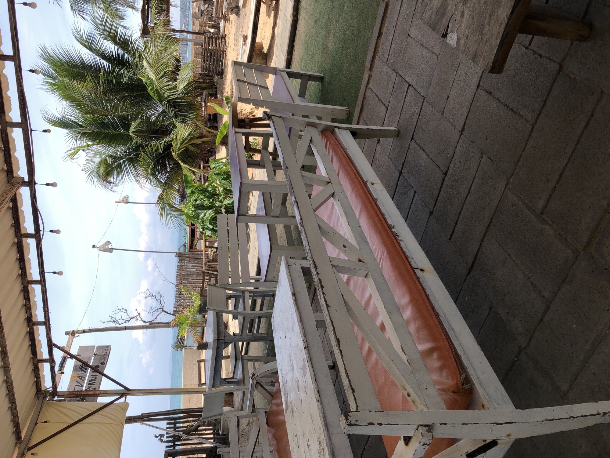 This is a photograph taken from a covered outdoor seating area overlooking the ocean in Mũi Né, Vietnam. The area features several wooden tables and chairs with white paint peeling, some with orange cushions. In the foreground, a long white bench with an orange cushion is visible. In the background, palm trees and the ocean can be seen. The sky is blue with some clouds. The scene suggests a beachside cafe or restaurant.