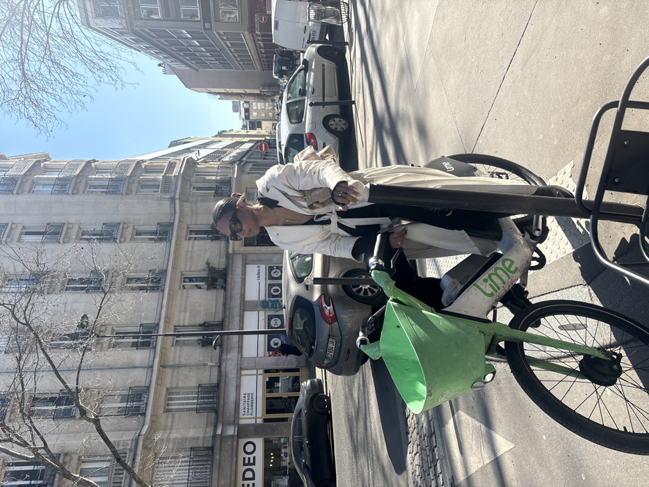 The image displays a street scene in Paris, France, on a clear, sunny day. A female individual is positioned centrally, stopped with a white and green Lime electric bicycle on a paved sidewalk. She wears a cream-colored trench coat over a black top, dark rectangular sunglasses, and a silver-colored pendant necklace, with her dark hair pulled back. Her left hand rests on the bicycle's handlebar, and her right hand holds onto a dark metal pole next to the bike. The Lime bicycle features a prominent bright green plastic basket at the front.
In the background, typical Parisian Haussmannian buildings, characterized by their light stone facade, multiple stories, numerous windows, and ornate iron balconies, line the street. At ground level, storefronts are visible, including one prominently displaying "CEDEO" and "SANITAIRE PLOMBERIE" (Sanitary Plumbing), confirming the French location. A street with parked and moving vehicles occupies the middle ground; a gray Peugeot 208 hatchback (French license plate CZ-694-JA) is clearly visible behind the bike. Bare branches of deciduous trees are seen against a clear blue sky on both sides of the frame. Strong sunlight casts distinct shadows on the ground and buildings.