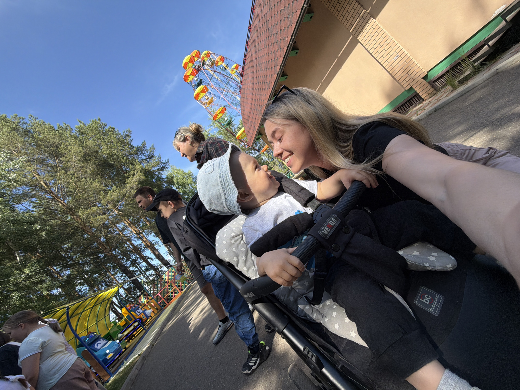 The image depicts an outdoor scene at an amusement park in Chaikovsky, Russia, under clear, sunny conditions. The scene is captured with a tilted perspective, dominated by a bright blue sky in the upper right.
In the foreground, a blonde-haired woman, wearing a black top and with sunglasses positioned on her head, smiles downwards and to her left. She is interacting with a young child seated in a dark-colored stroller immediately in front of her. The child, wearing a light blue bucket hat and a grey t-shirt, reciprocates the smile and looks up at the woman. A grey blanket with white star patterns covers the child's legs within the stroller. The stroller features "VE GA" branding on its handlebar and a "bc Babycare" logo on its footrest.
Behind the woman and child, several individuals are visible. A man wearing a black cap and black shirt is partially obscured by the stroller. Behind him, another man in a black t-shirt, blue jeans, and black athletic shoes is walking. Further to the right in the mid-ground, a blonde-haired woman in a plaid shirt looks upward and to her right. On the far left, a young girl in a light-colored t-shirt stands near an amusement park ride.
The background features several elements. To the left, a colorful amusement ride with a yellow canopy and multi-colored vehicles (blue, yellow, red) is visible. To the right, a large Ferris wheel with distinct yellow, orange, red, and green cabins stands prominently against the blue sky. Adjacent to the Ferris wheel, a building with a reddish-brown tiled roof and light brown/tan walls, detailed with a green trim and a brick-patterned section, is present. Tall green trees with dense foliage are visible across the upper left and top edges of the image. The ground consists of a dark, paved surface, illuminated by sunlight.