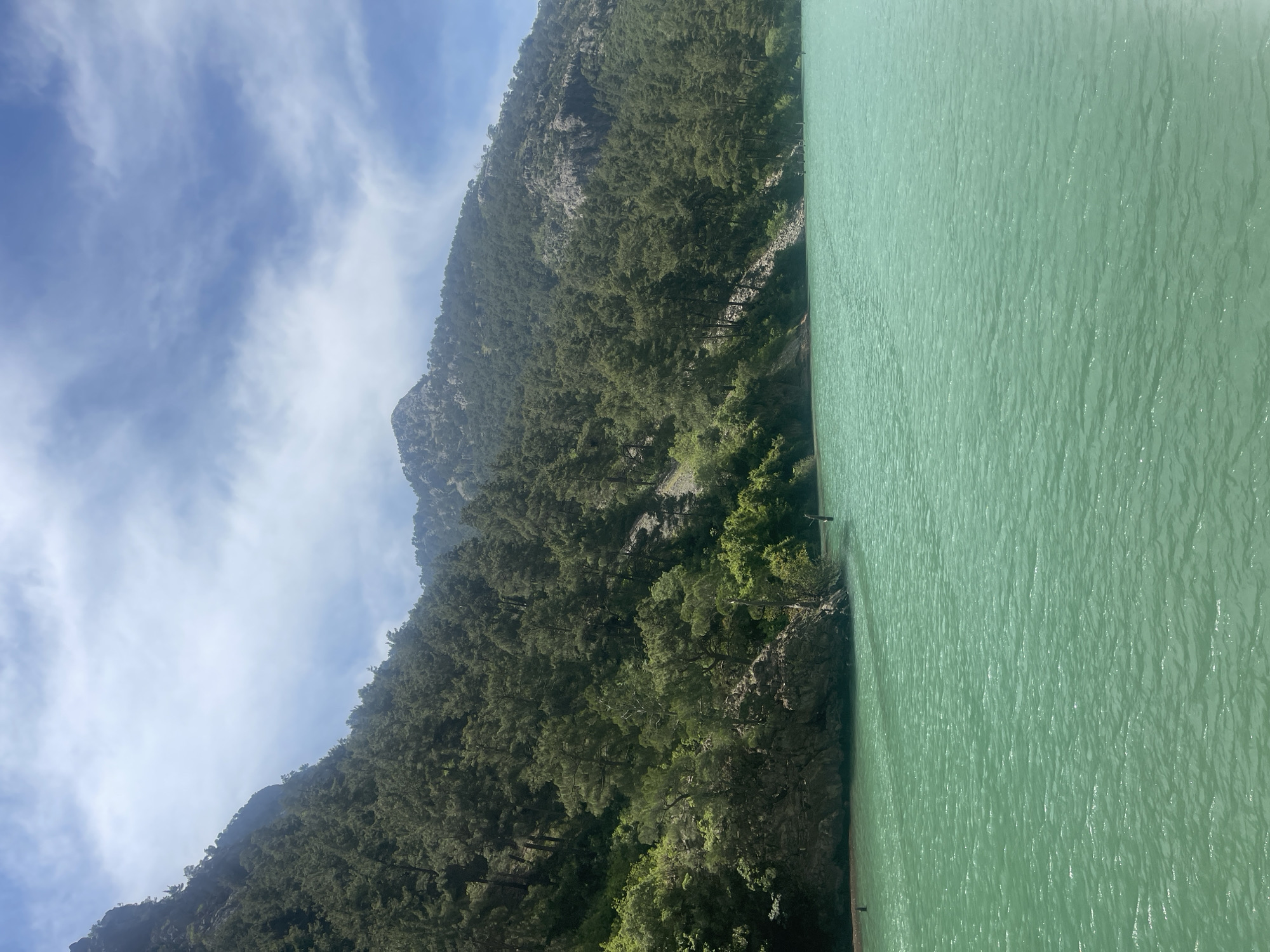 The image presents a scene of a body of water bordered by a mountain densely covered with trees. The water's color is a pale green. The sky is blue with scattered clouds. The location is Unknown, Unknown.