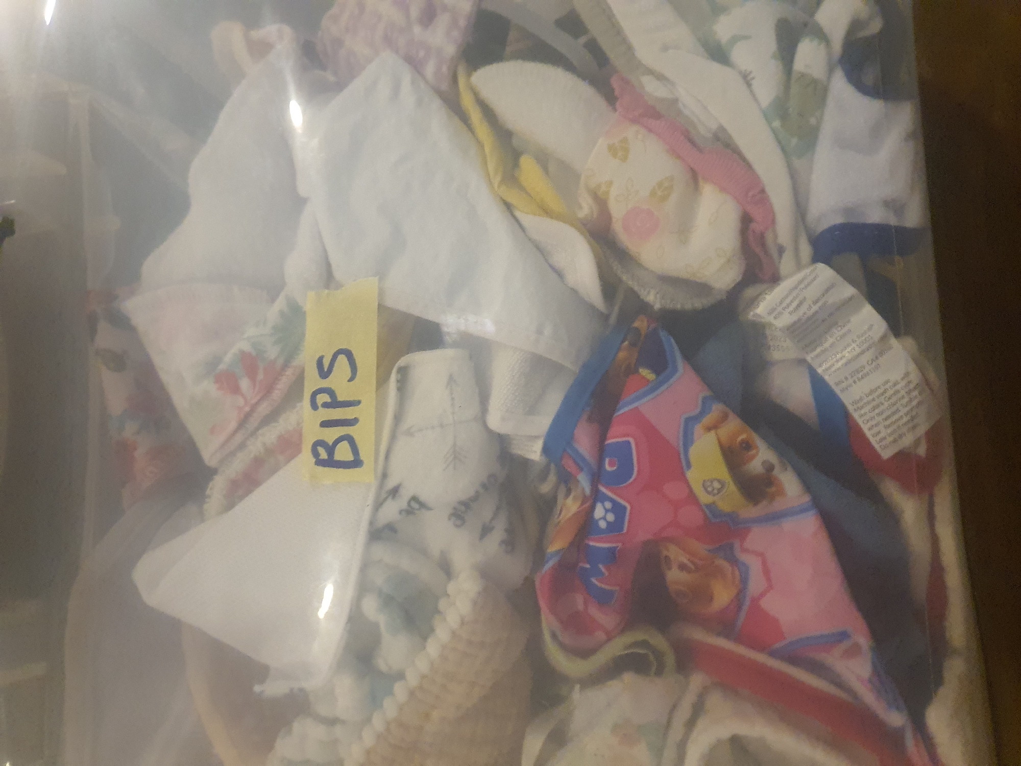 The image shows a close-up view of a clear plastic bin filled with various baby bibs, or "BIPS" as indicated by a yellow sticky note with blue writing. The bibs are made of different fabrics and feature a variety of patterns, including floral designs, geometric shapes with arrows, and characters from the Paw Patrol cartoon. Some bibs are white, while others have pink, yellow, green, and blue accents. The lighting in the image is bright, creating some glare on the plastic bin. The background is dark and out of focus, with a hint of what appears to be a wooden surface. The overall impression is one of organized storage for baby clothing items. There is also a garment care tag visible in the lower right portion of the image, containing washing instructions. The word "BIPS" is the only clearly legible text.