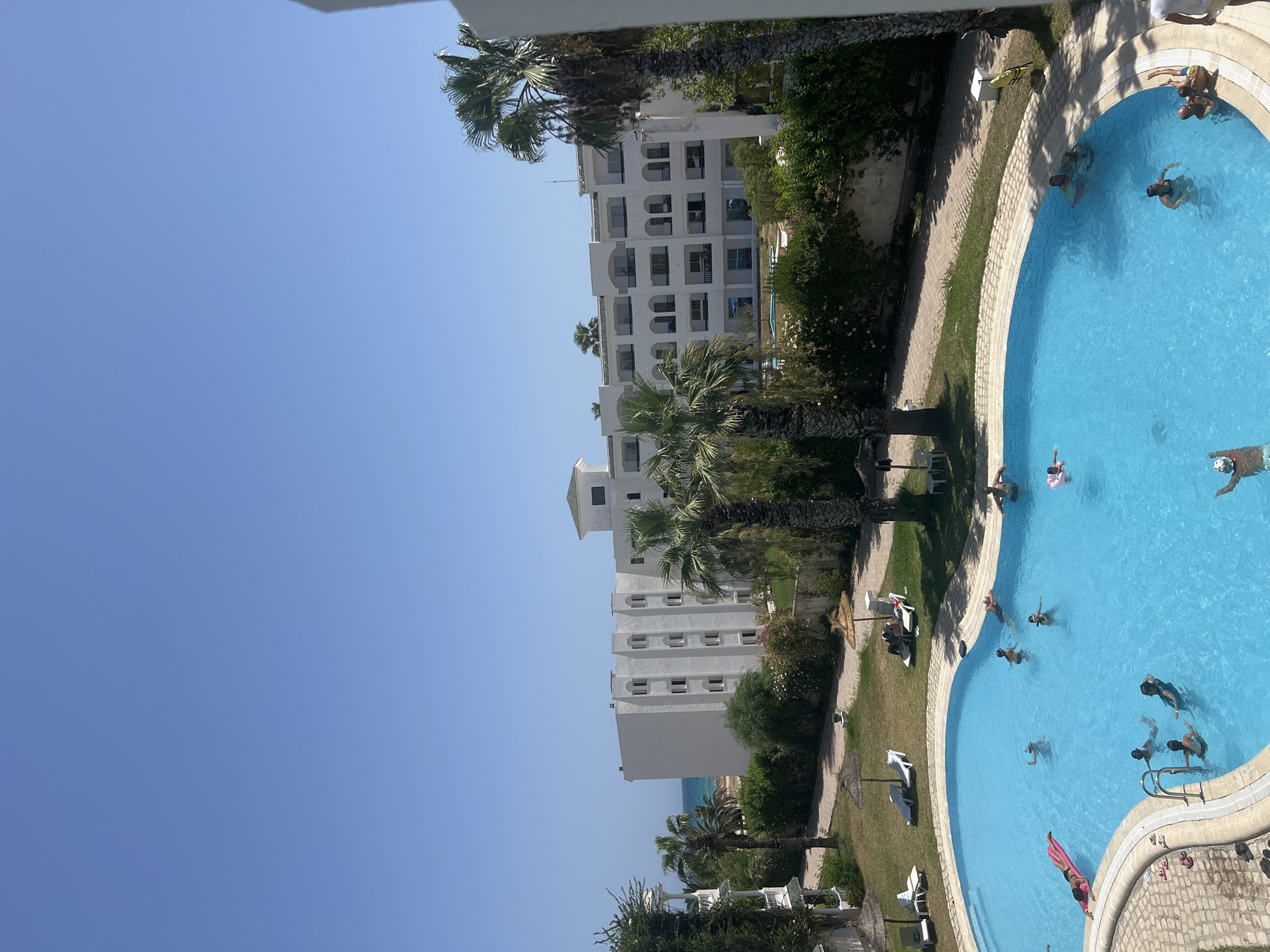 The image shows an outdoor swimming pool at a resort in Hiboun, Tunisia. The pool is filled with clear blue water and several people are swimming and relaxing in it. There is a pink inflatable raft in the pool, and a woman is lying on it. There are also lounge chairs and umbrellas around the pool, with some people sunbathing on them. In the background, there is a white hotel building with balconies, palm trees, and lush green vegetation. The sky is clear and blue, indicating a sunny day. The scene depicts a typical holiday resort setting.