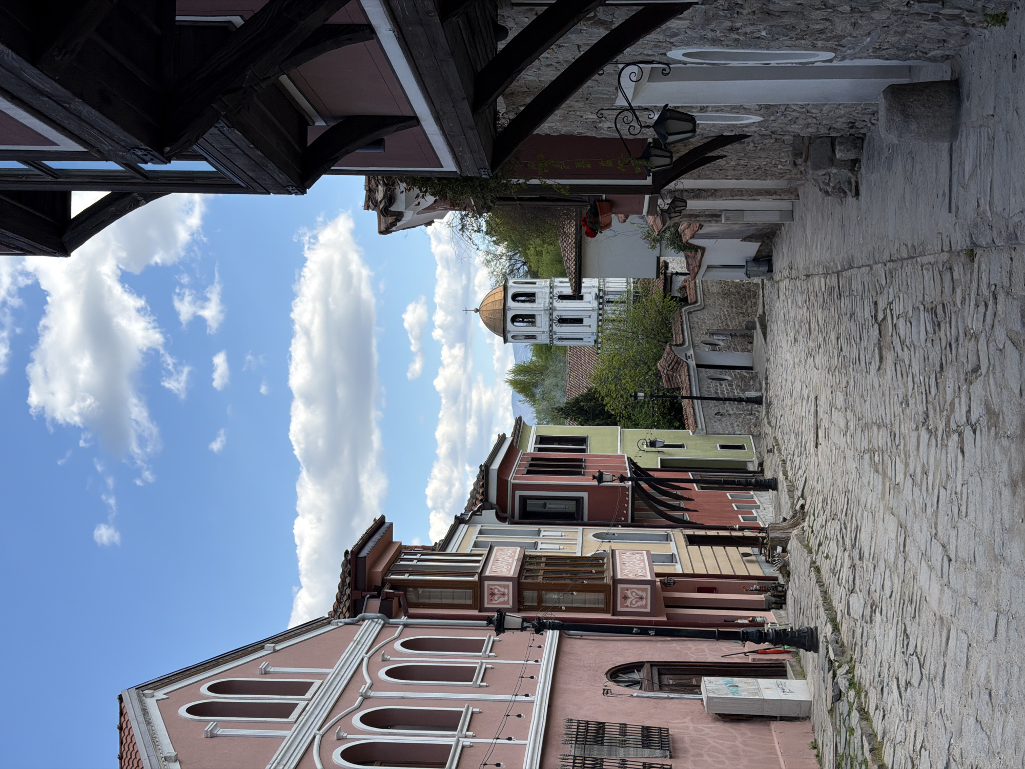 This media file captures a picturesque cobblestone street winding through the historic Old Town of Plovdiv, Bulgaria, under a bright blue sky filled with scattered white cumulus clouds. The scene is illuminated by clear daylight, suggesting a pleasant day.
The narrow street, paved with irregular grey cobblestones, recedes into the distance, flanked by traditional multi-story houses on both sides. On the left, a prominent pink/peach building in the foreground showcases distinctive architectural elements, including arched windows, white decorative trim, and an ornate facade with intricate mouldings. A partial word, "BULG," is visible on a white sign or box attached to this building. Further down the street on the left, a row of colorful houses (red, yellow, light green) display characteristic Bulgarian National Revival architecture, featuring prominent dark wooden beams, overhanging upper floors, and ornate bay windows with intricate patterns.
On the right side of the street, the upper section of a building with dark wooden structural elements is visible, possibly forming an overhang. Ornate street lamps are attached to several building facades, adding to the historic ambiance. In the mid-ground, nestled between the buildings and lush green trees, stands a white church bell tower topped with a golden dome and a cross. The tower features multiple arched openings. No people or specific activities are visible, conveying a sense of quiet charm and historical preservation.
