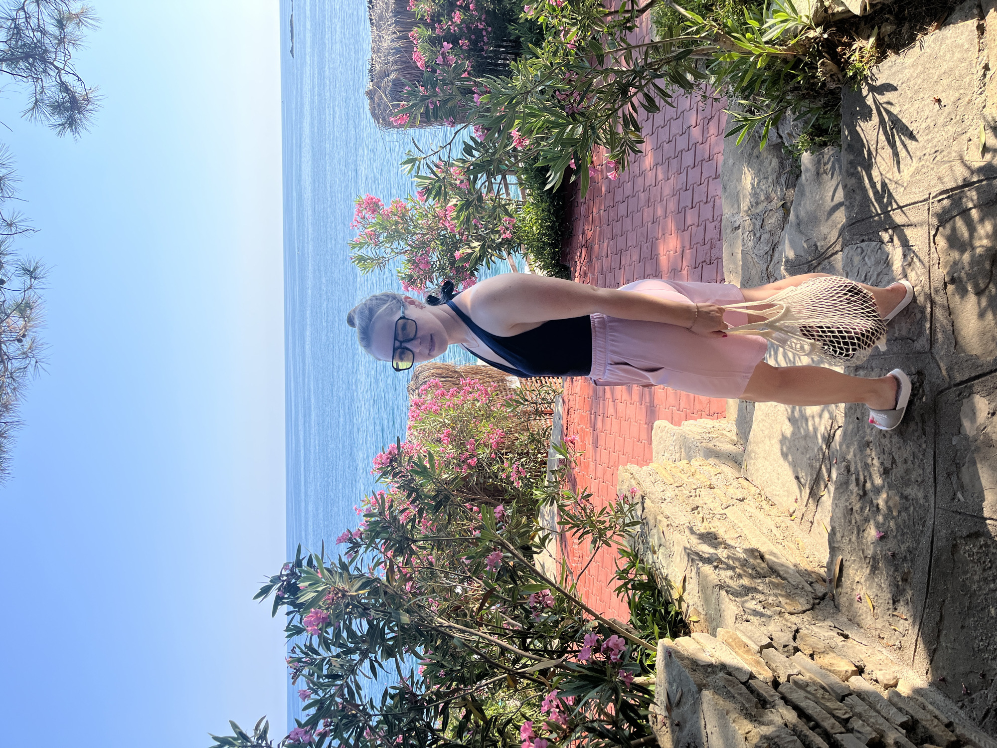 A young Caucasian woman with blonde hair pulled into a bun, wearing glasses, a black one-piece swimsuit, and light pink shorts, stands on a stone pathway next to a wall. She is carrying a white mesh bag with dark items inside. She is positioned on a stepped pathway that leads down towards a red brick patio area. Behind her, vibrant green bushes with pink flowers are in bloom. The background features a clear blue sky and the tranquil blue sea. This scene is located in Alanya, Turkey.