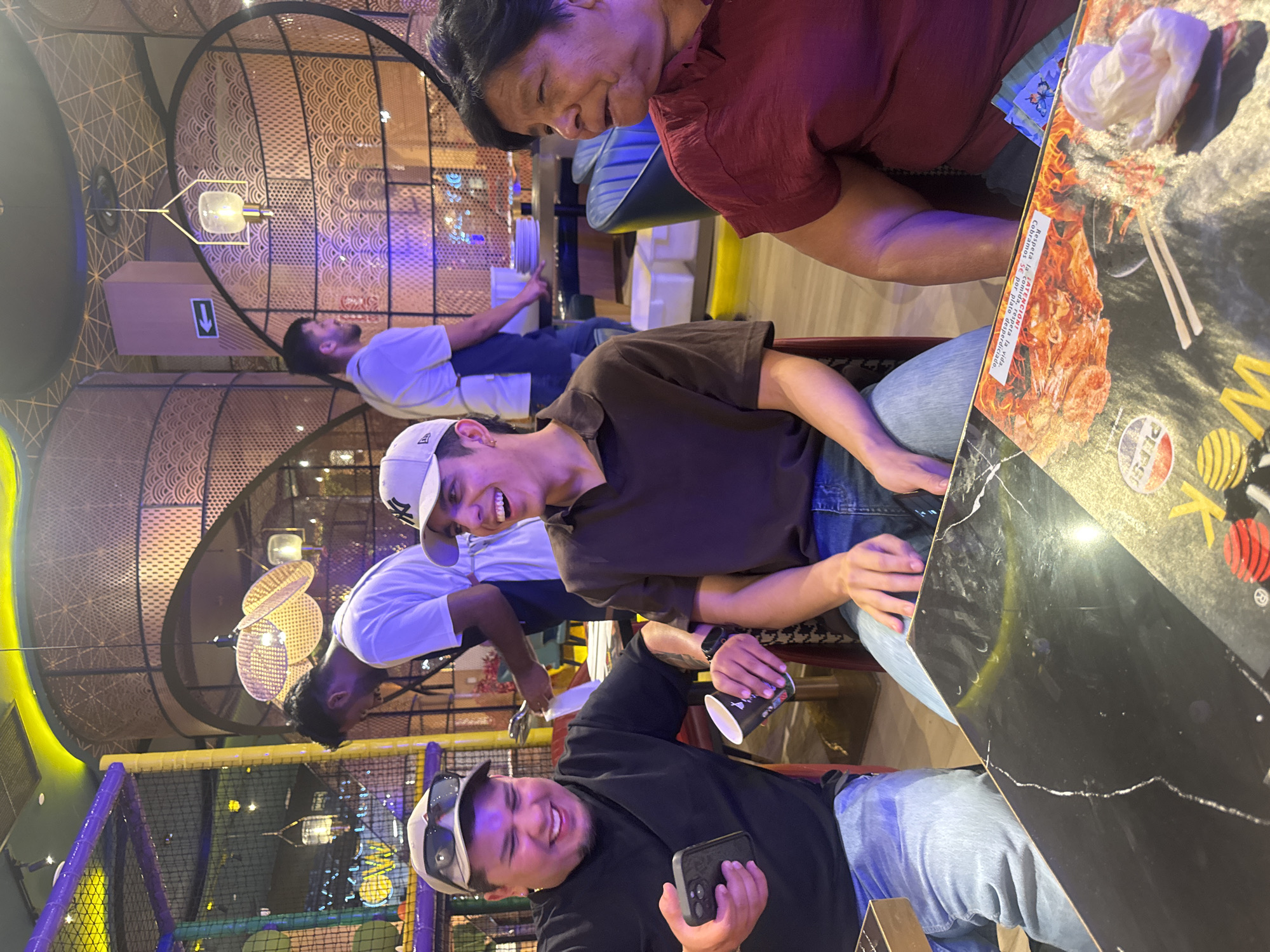 The image shows a group of people sitting at a table in what appears to be a restaurant or entertainment venue. The setting features modern decor with circular patterns and ambient lighting. The people are casually dressed and seem to be enjoying themselves.
In the foreground, two men are visible. One is wearing a black long-sleeved shirt, a baseball cap, and sunglasses pushed up on his head. He is smiling and holding a smartphone. Next to him, another man in a brown t-shirt and a white baseball cap is also smiling, looking towards the camera. Behind them, a woman in a red shirt and an older person, partially visible, are also seated at the table. Another person in a light blue t-shirt is seated further back.
The table surface is black and reflective, with a menu or placemat featuring an image of food and some text. The visible text on the placemat includes "K", "PEPSO", and phrases in Spanish that seem to relate to respecting food and a charge for wasted plates. The presence of a playful, brightly colored play area in the background, with a net structure, suggests the location might be a family-friendly venue or a restaurant with an integrated play area. The lighting suggests it could be evening or indoors with artificial lighting. The overall mood appears relaxed and convivial.