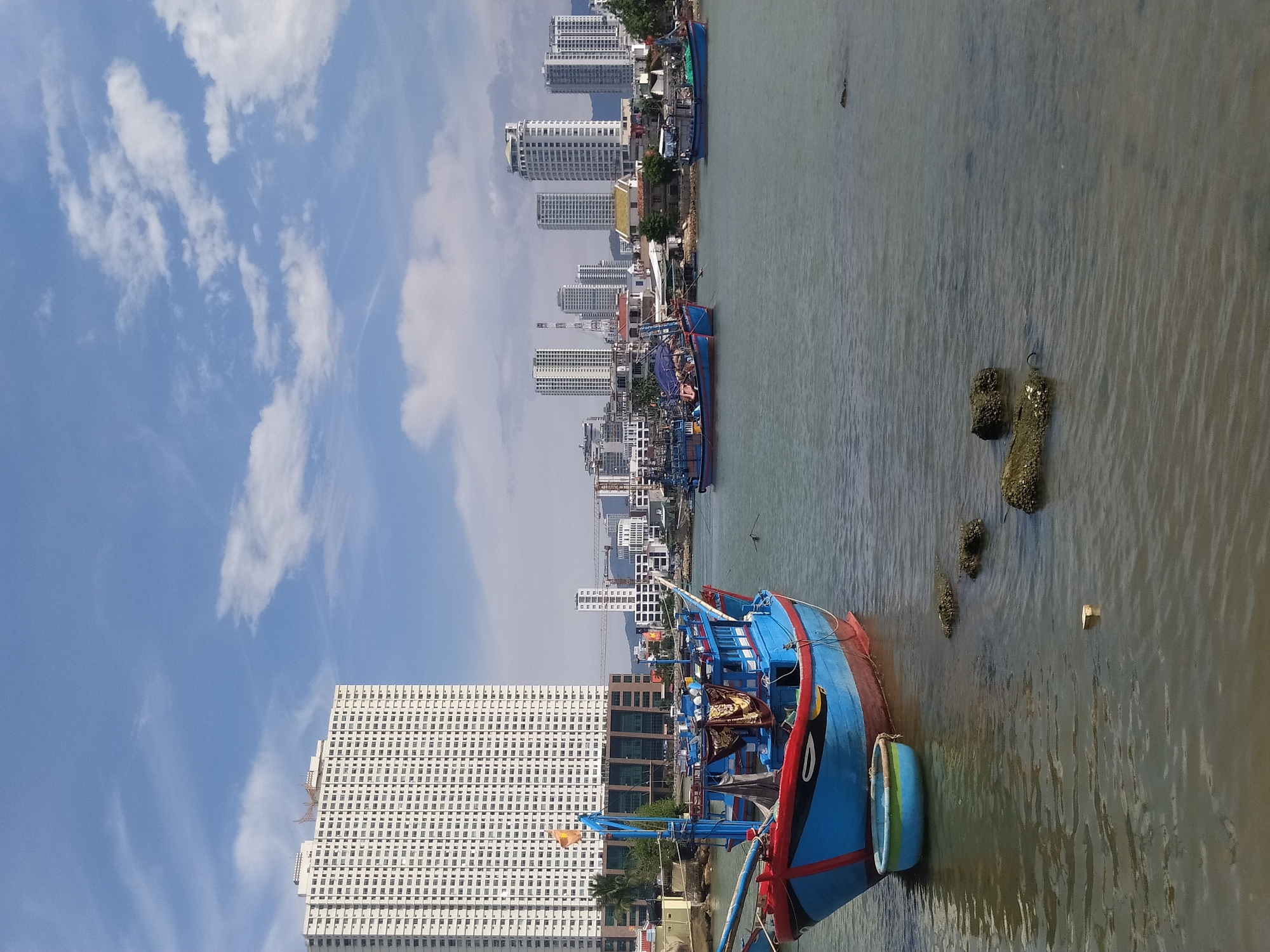 An eye-level, medium shot captures a harbor scene with numerous buildings and boats under a bright, partly cloudy sky. In the foreground, a vibrant blue and red fishing boat is docked, with a small, blue, bowl-shaped boat tied to its side. Several rocks protrude from the murky water near the boat. In the background, a cluster of modern, tall buildings dominates the skyline, with a very tall, white residential building on the left side of the frame. Other smaller buildings and cranes are visible between the larger structures. Several other fishing boats are scattered across the water. The overall impression is of a busy harbor with both traditional maritime elements and modern urban development.
