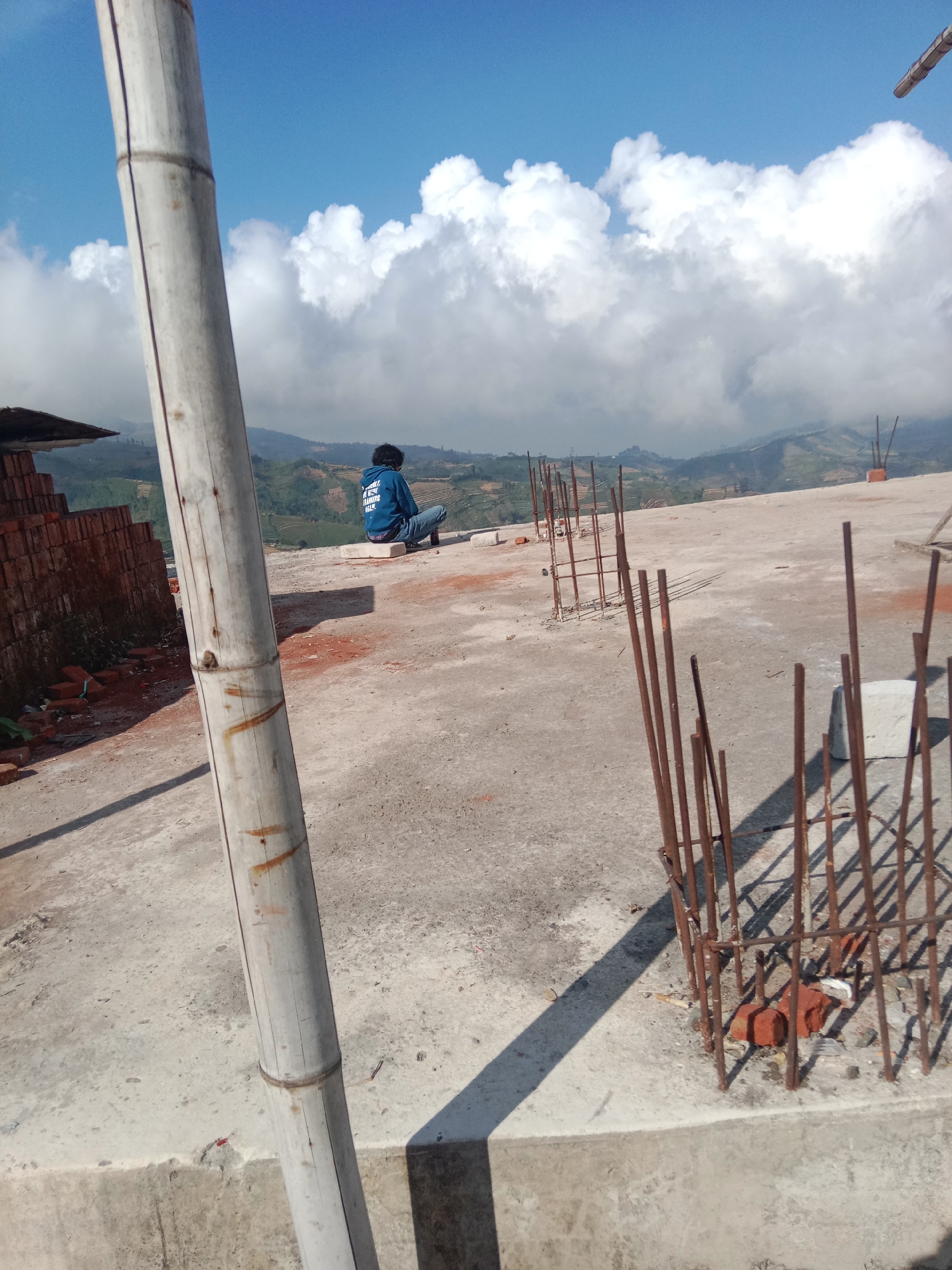 The image depicts a rooftop construction site overlooking a hilly, rural landscape with terraced fields under a bright, cloudy sky. A person wearing a blue hooded jacket and jeans sits on a white concrete block, facing away from the camera and towards the scenic view. The rooftop is made of concrete and shows signs of ongoing construction, with piles of bricks, rebar protruding from the ground, and a large bamboo pole positioned vertically in the foreground. The overall impression is one of a paused moment of contemplation amidst the work, with the natural beauty of the surrounding area providing a striking backdrop. The location is identified as Tieng, Indonesia.