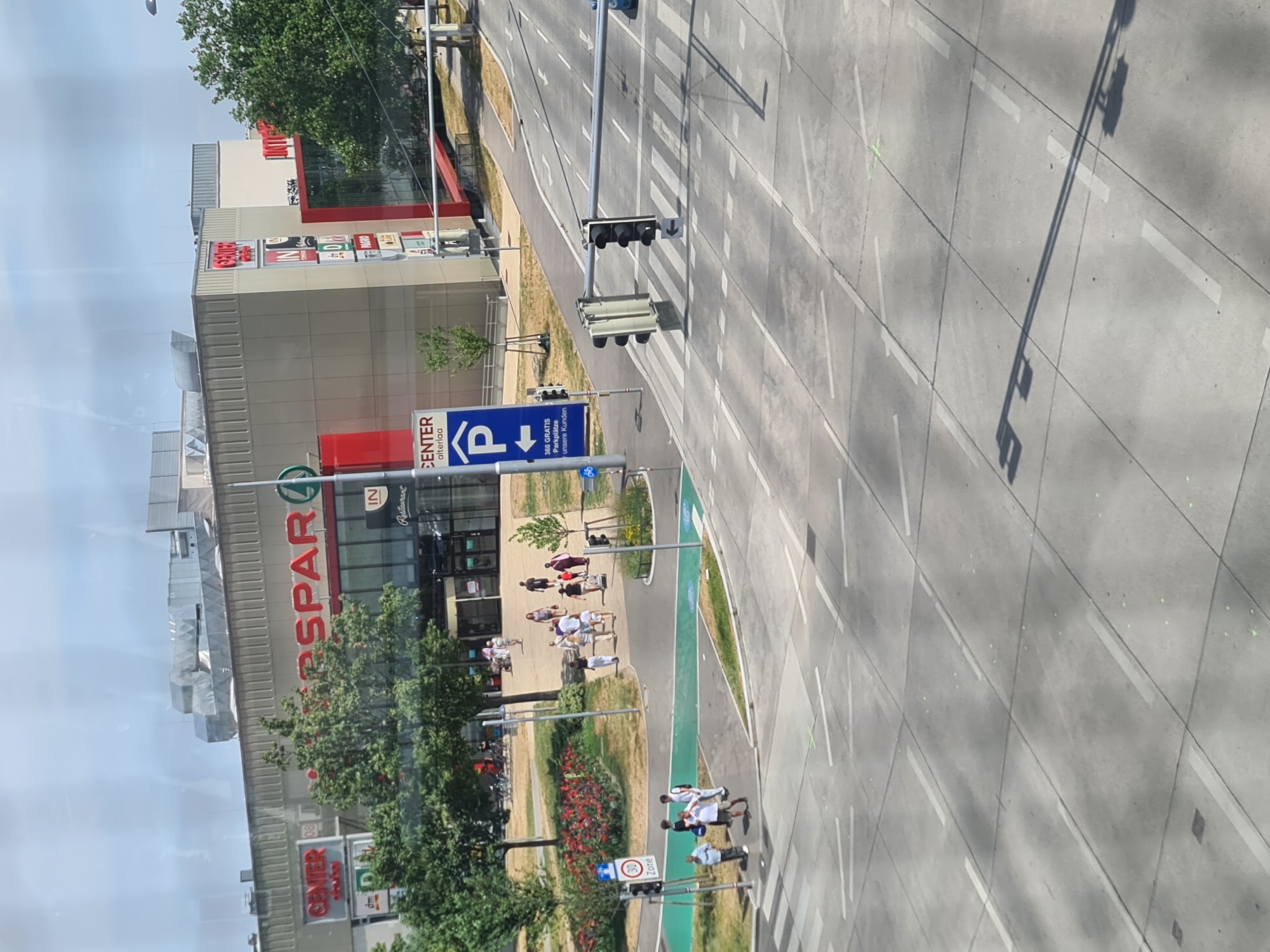 The image captures a street view in Vienna, Austria, likely near a shopping center called "CENTER". In the foreground, a street with crosswalks and traffic lights is visible. Several vehicles, including a blue car, are present on the road. Pedestrians are seen crossing the street and walking on the sidewalk.
The background features a multi-story building with various retail signs, including "SPAR" and "dm". There are also trees and greenery around the building. The sky is bright and cloudy, indicating daylight. The overall scene depicts a typical urban environment with commercial activity and pedestrian traffic. The presence of German signage suggests the location is in a German-speaking region.