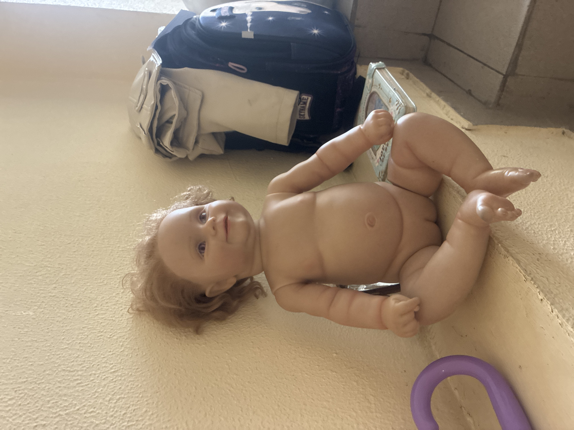 A lifelike baby doll is lying on its back, positioned on a textured, light-colored surface that appears to be a wall or floor. The doll is naked, with soft, realistic skin, blonde curly hair, and purple eyes. Its arms and legs are bent in a playful pose.
Behind the doll, a dark blue backpack with a starry night pattern and a white and tan garment is visible. To the right of the doll, a decorative box with a floral pattern and the words "Love" are also present. A small purple toy, possibly a ring or handle, is in the lower right corner of the image.
The scene suggests a child's play area or a storage space. The lighting appears to be natural, casting soft shadows. No specific time of day or weather conditions are discernible. The context of Sochi, Russia, doesn't provide further visual cues within the image itself.
The text "SKINAME" is visible on a label attached to the white and tan garment.