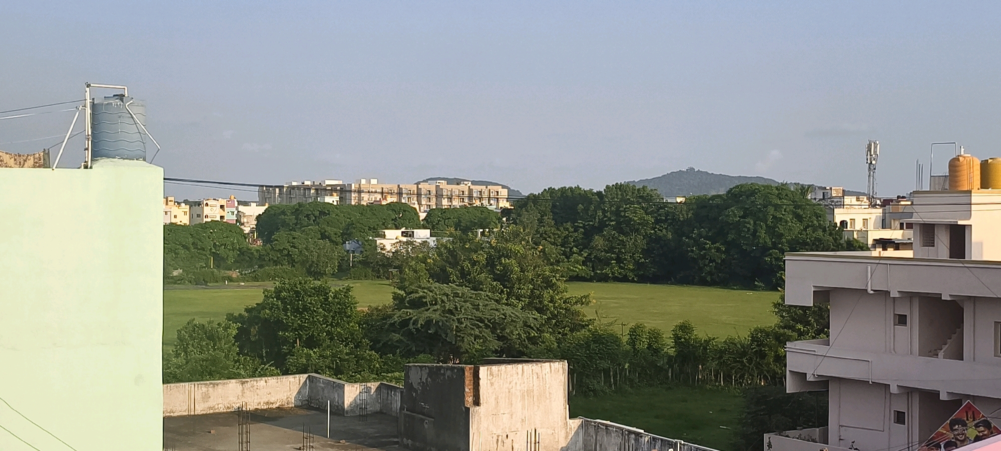 The image shows a panoramic view of a city with buildings, trees, and a grassy field. In the foreground, there are rooftops of buildings. A large light green building is on the left, and a white building with multiple balconies is on the right. A water tank is visible on top of the green building. In the midground, there are lush green trees and a vast open grassy field, possibly a park or a sports ground. Beyond the trees, several multi-story buildings and a cell tower can be seen. In the background, a mountain range is visible under a clear sky. The scene appears to be a suburban or semi-urban area of Kundrathur, India, with a mix of residential buildings and natural landscapes. The lighting suggests it is daytime, possibly late afternoon due to the warm tones. There are no people or animals visible in the frame.