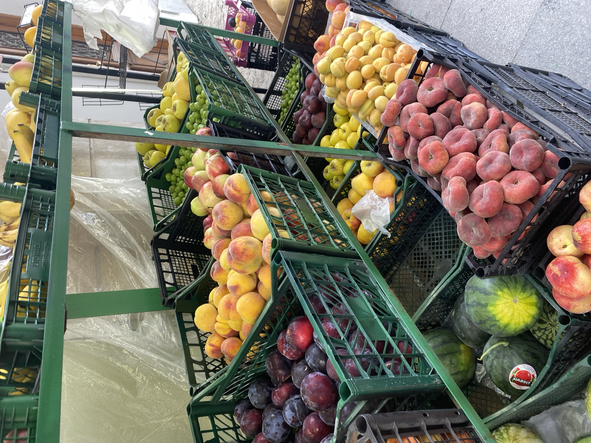 This is a medium-shot, eye-level photograph of a fruit stand in Batumi, Georgia. The stand is made of green metal shelves holding several plastic crates filled with various fruits. From top to bottom, left to right, the crates contain bananas, lemons, apples, plums, nectarines, apricots, peaches, and watermelons. The fruit stand is set up outdoors, with a translucent plastic sheeting visible behind the fruits, suggesting it might be a covered market or stall. The ground is paved. There are no people visible in the image, and the fruits are the sole focus of the scene. The overall impression is one of abundance and freshness.