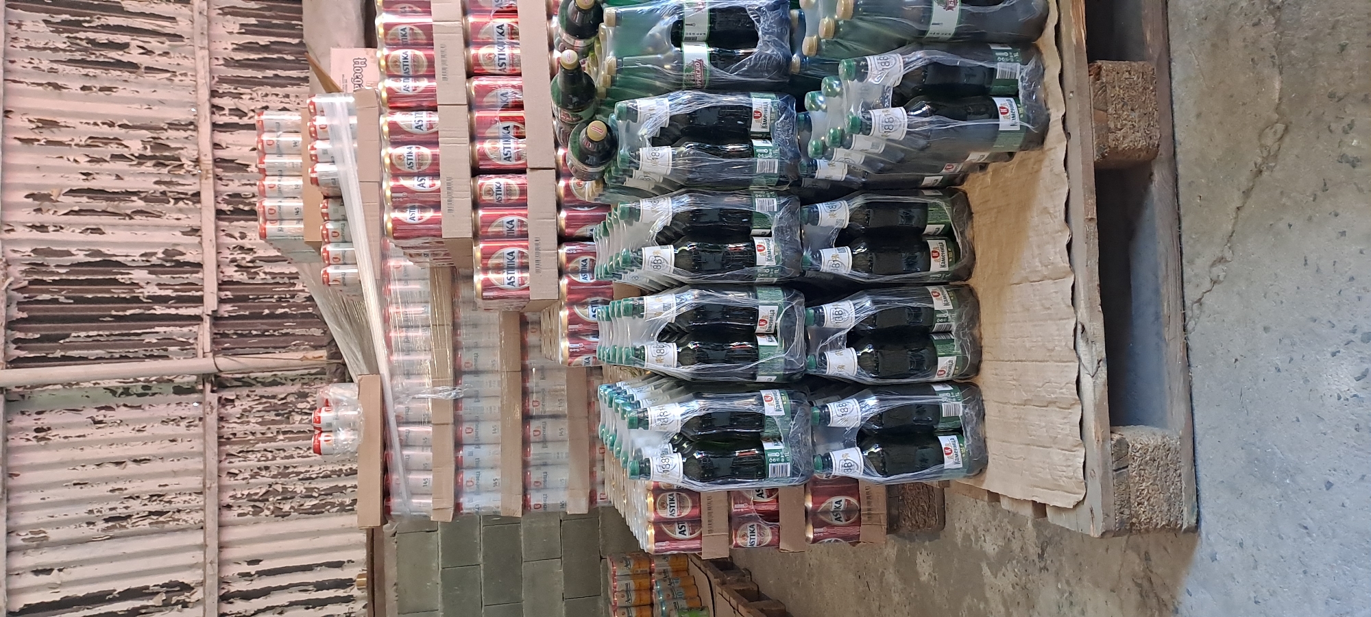 The image shows a storage area filled with crates and pallets of bottled and canned beverages. The scene appears to be an indoor warehouse with a corrugated metal ceiling that is peeling and weathered. The floor is concrete.
In the foreground and middle ground, there are multiple stacks of bottled beverages, predominantly dark glass bottles packaged in plastic shrink wrap. Many of these bottles have green caps and labels with Cyrillic text, including the brand name "КАМЕНИЦА" and "1881". Some bottles are also visible in the background, with a few green and yellow cans also present.
Further back, numerous stacks of canned beverages are visible, also packaged in cardboard boxes and shrink wrap. These cans are primarily red and white, with some labels showing the brand "ASTIKA". There are also stacks of what appear to be larger cylindrical containers, possibly larger cans or containers of other beverages. The arrangement of the products suggests an organized inventory system.
The overall impression is of a commercial storage facility for beverages, likely a distribution center or warehouse. The context of Velika, Bulgaria, suggests this location is a specific commercial establishment within that region.