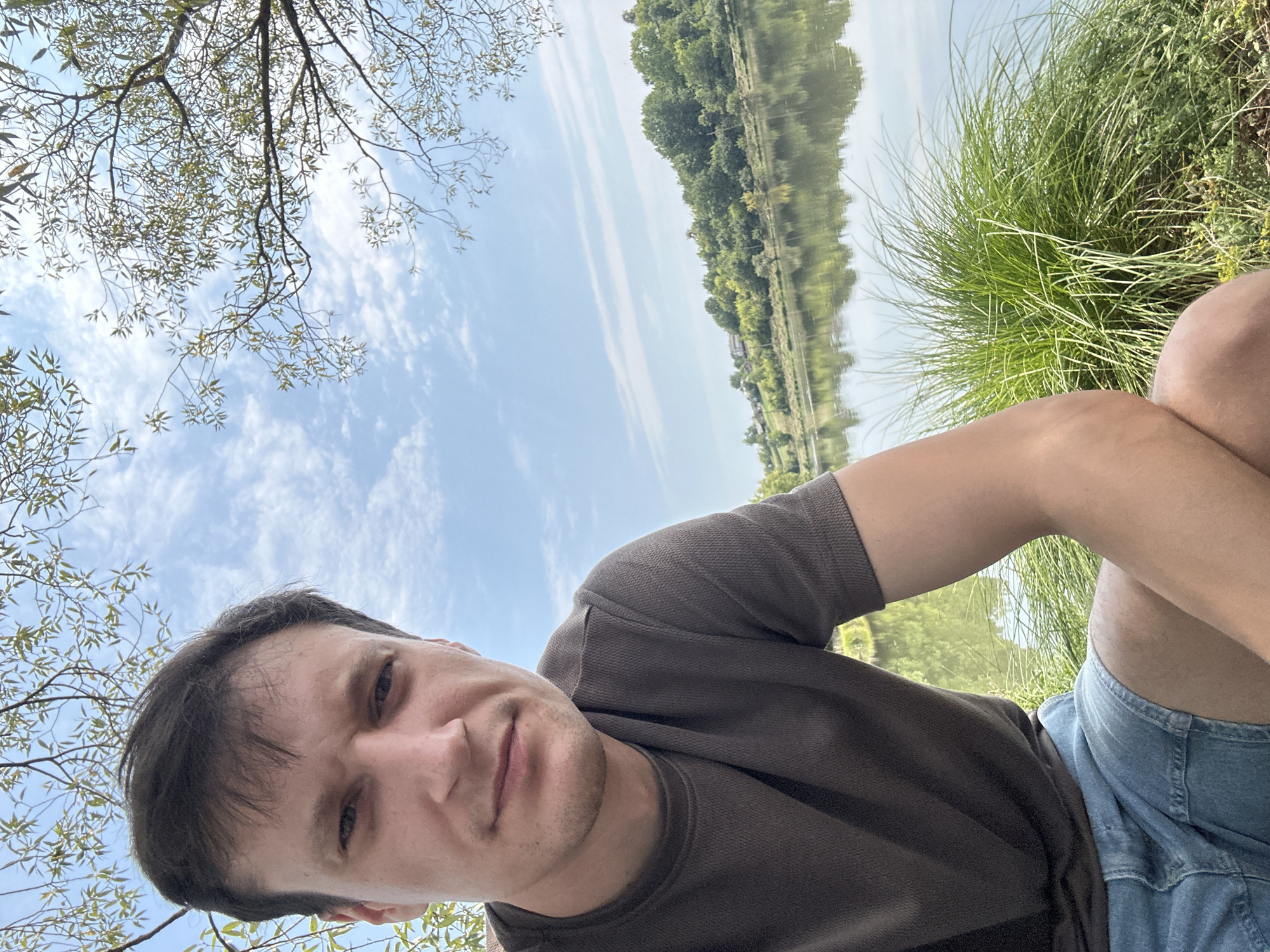 This is a selfie taken by a man with short, dark brown hair and a fair complexion. He is wearing a dark grey, textured t-shirt and light blue denim shorts. The man is positioned in the foreground, looking towards the camera with a neutral expression. Behind him, a body of water, likely a river or lake, is visible, reflecting the blue sky and scattered white clouds. The opposite bank is lined with lush green trees and some scattered houses, suggesting a rural or semi-rural setting. Tall green grass and some foliage are in the immediate foreground to the right of the man. The overall scene depicts a pleasant outdoor setting on a clear day.