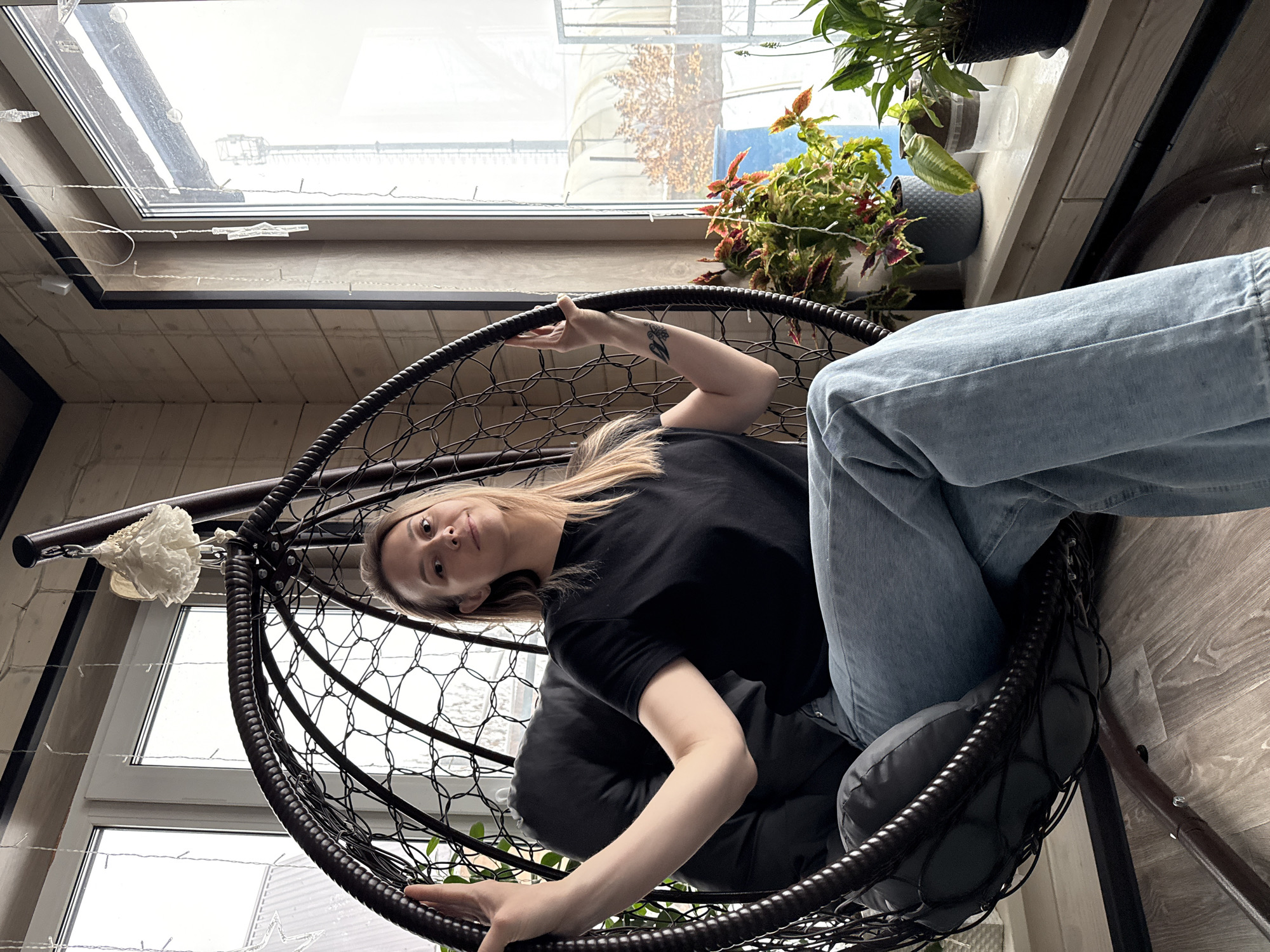 A woman with blonde hair is sitting in a dark-colored, hanging wicker chair indoors, looking at the camera. She is wearing a black t-shirt and blue jeans. Her left arm is extended, holding the top of the chair's frame. A tattoo is visible on her left forearm. The chair has a gray cushion.
Behind the woman, a window shows an exterior view, possibly a building or structure. There are plants in pots on the windowsill. String lights are visible near the window frame. The flooring appears to be wooden.
The scene is an interior shot, seemingly in a room with natural light coming through the window. The city of Chaikovsky, Russia, is not explicitly indicated in the image. However, it might be inferred based on metadata or external information.