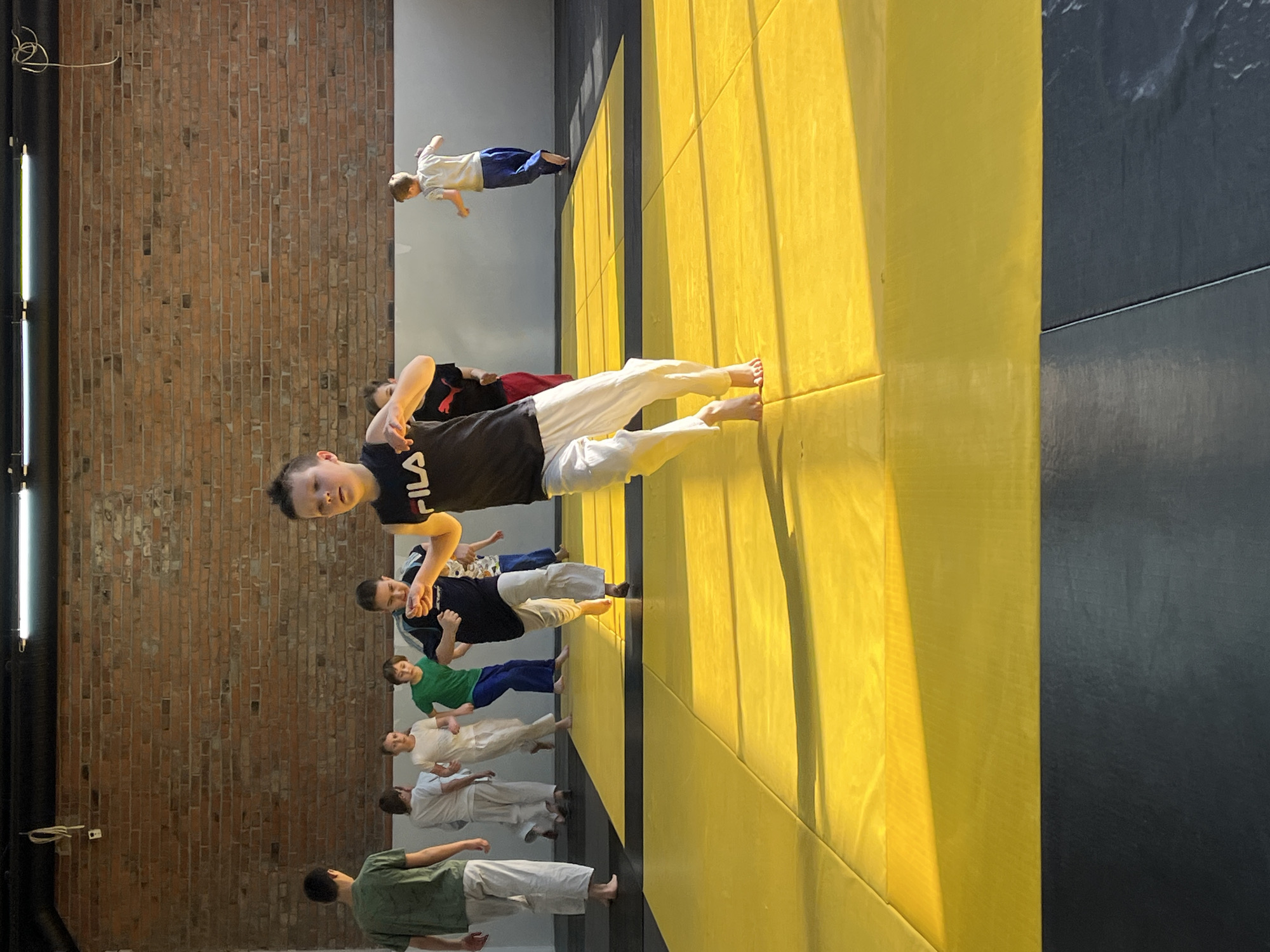 A group of children and teenagers are engaged in a martial arts class in a large training room. They are standing on a yellow mat that is adjacent to a black mat, and in a line near a brick wall. One child is wearing a black shirt, white pants, and is facing the camera and smiling. Other children in the line are wearing various combinations of white pants and shirts, green shirts and blue pants, and black shirts and maroon pants. The scene is lit by natural light and a bright fluorescent light fixture on the left side of the image. The location is presumably in Krasnoyarsk, Russia.