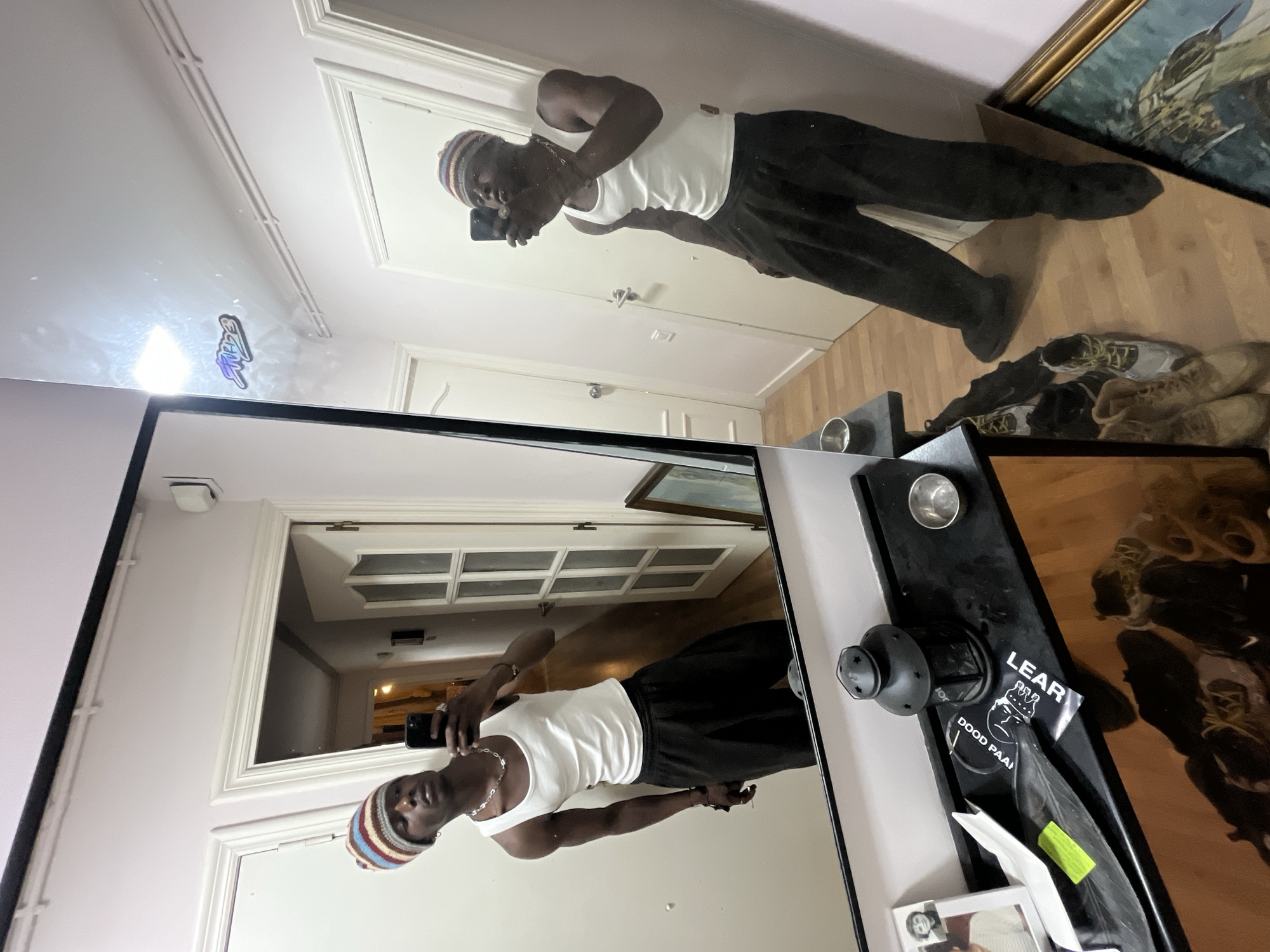 A person is taking a selfie in a mirror. The person is wearing a white tank top, black pants, and a striped beanie. The mirror is reflecting a room with a door, a hallway, and a dresser. The dresser has various items on it, including shoes, a picture frame, and some decorative objects. The location is undetermined.