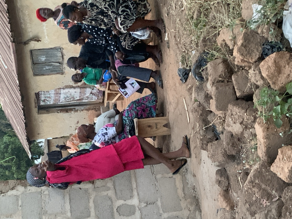 A group of people are seated outdoors, some on chairs and others on the ground. They appear to be gathered for a meeting or event next to a building with a corrugated metal roof. A woman in a bright pink dress is in the foreground. The ground is dirt and some bricks are visible to the right. Several people are holding papers, suggesting they are following along with a presentation.