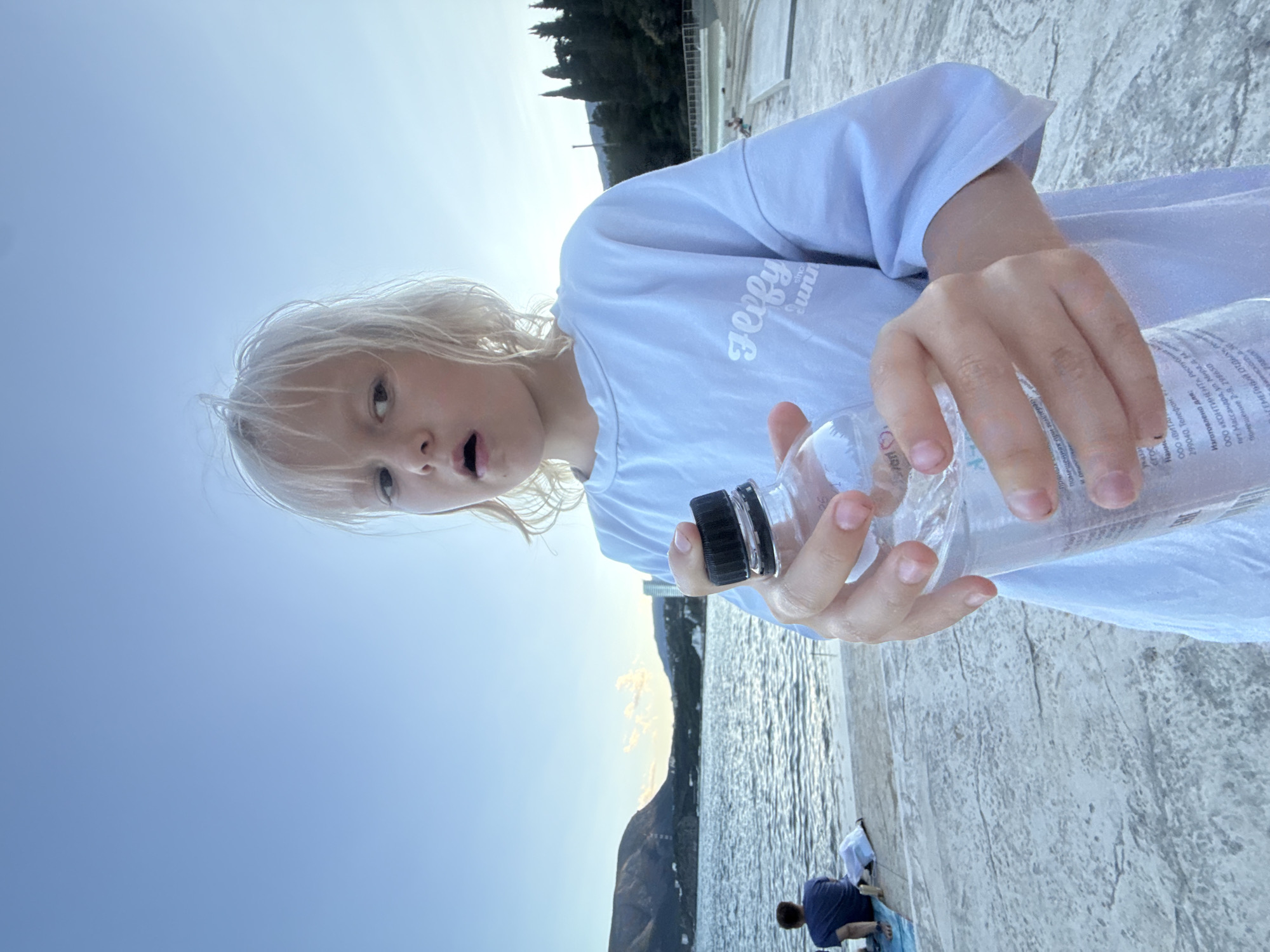 A young child with blonde hair stands on a concrete walkway near a body of water, holding a clear plastic bottle of water. The child is wearing a light blue t-shirt with a white graphic design. In the background, there is a person sitting on a blue mat by the water, and beyond that, hills and trees under a clear sky. The scene appears to be a waterfront area, possibly a promenade or park.
