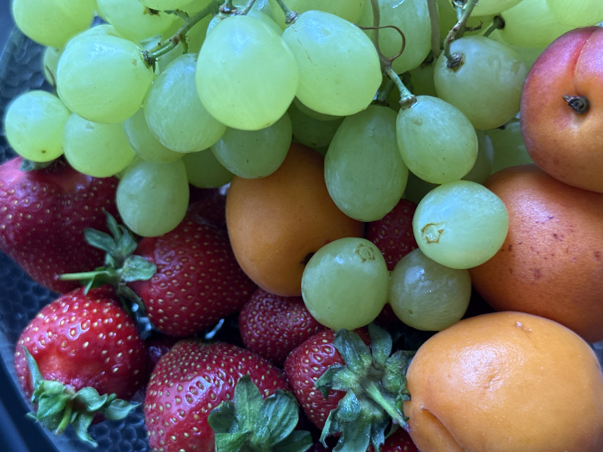 The image displays a close-up, overhead view of a bowl filled with various fruits. Prominently featured are bright red strawberries with visible seeds and green leafy tops, a cluster of pale green grapes, and several orange-hued fruits, likely apricots or peaches, with a smooth skin. The fruits are closely packed together, creating a vibrant and colorful arrangement. The lighting suggests it is daytime, and the background is dark and out of focus, likely the interior of a bowl or container. There is no identifiable location in the city of Krasnoyarsk, Russia, or any other specific geographic information present within the image itself. The scene is static, with no visible actions or interactions occurring.