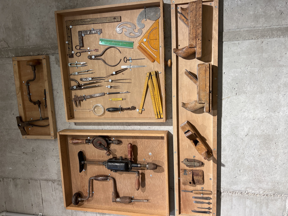 This image depicts a collection of vintage hand tools, primarily woodworking and drafting instruments, meticulously displayed on a concrete wall in the city of Yokneam Ilit, Israel. The tools are organized within four separate wooden-framed display units, each featuring a light-colored plywood backboard to which the items are secured.
The topmost rectangular display contains a large adjustable wrench and a brace drill. Below this, on the left, a larger vertical display box holds three distinct hand drills: two brace drills and one eggbeater-style hand drill. To its right, another large display box exhibits a diverse array of measuring and drafting tools, including a large calipers, multiple compasses and dividers, a pair of scissors, a metal set square, a green plastic ruler, an orange plastic protractor, a metal French curve, a yellow folding rule, a magnifying glass, and various scribing and marking implements. The bottom-most long horizontal panel displays a row of smaller hand tools such as chisels, a small saw blade, a utility knife, two leather tool pouches, and four different wooden hand planes.
Many of the tools, particularly those made of metal, exhibit visible signs of age, wear, and surface oxidation, consistent with historical use or preservation. All items are individually fastened to their respective backboards, indicating an exhibition or educational arrangement.