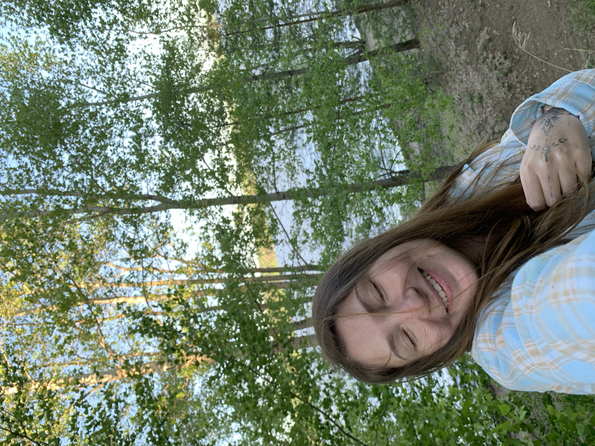 The image is an eye-level selfie, vertically oriented, taken outdoors. The subject is a smiling female with long brown hair, wearing a light blue and tan plaid shirt. Her right hand, adorned with a geometric tattoo, is touching her hair. The background is a natural scene including trees, green foliage, and a body of water. It is not possible to identify a precise location in the city of Chaikovsky, Russia without more specific information.