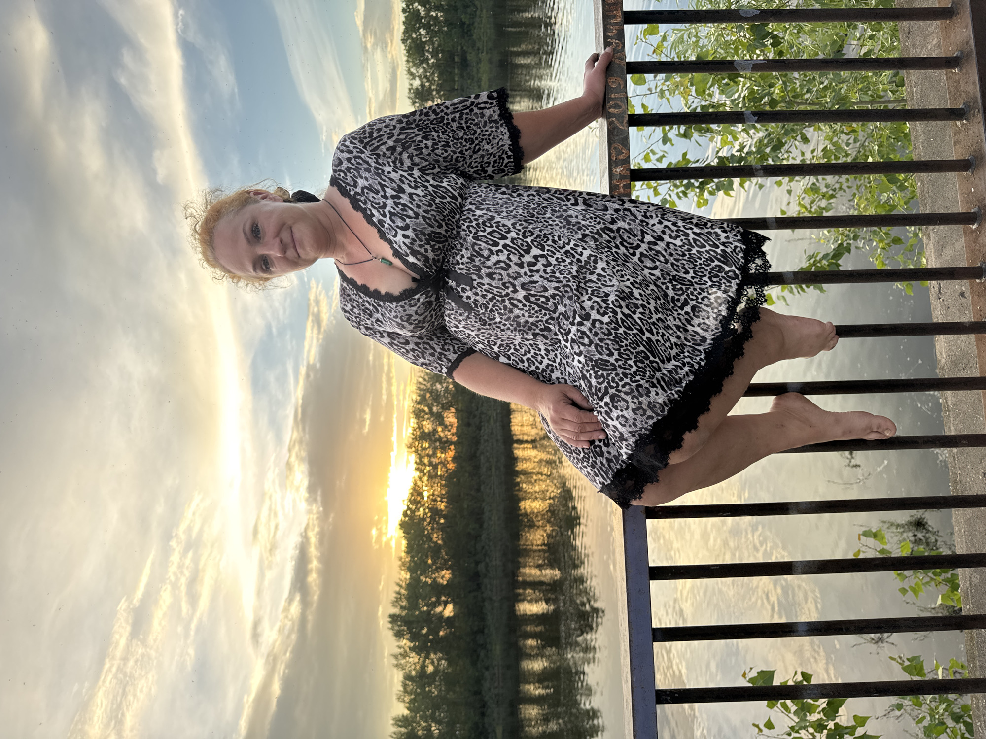The image shows a woman with blonde, curly hair wearing a leopard-print dress and sitting on a metal railing. She is looking at the camera and smiling. Behind her, a river is visible with trees on the opposite bank, and the sunset is setting in the distance, casting a warm glow on the sky and water. The railing is dark metal and has some graffiti on it. The woman's right hand is resting on the railing, and her left arm is bent. Her legs are dangling over the railing, and her bare feet are visible. The scene appears to be a public walkway or bridge overlooking the Kama River in Chaikovsky, Russia, given the distinctive landscape and the bridge railing.