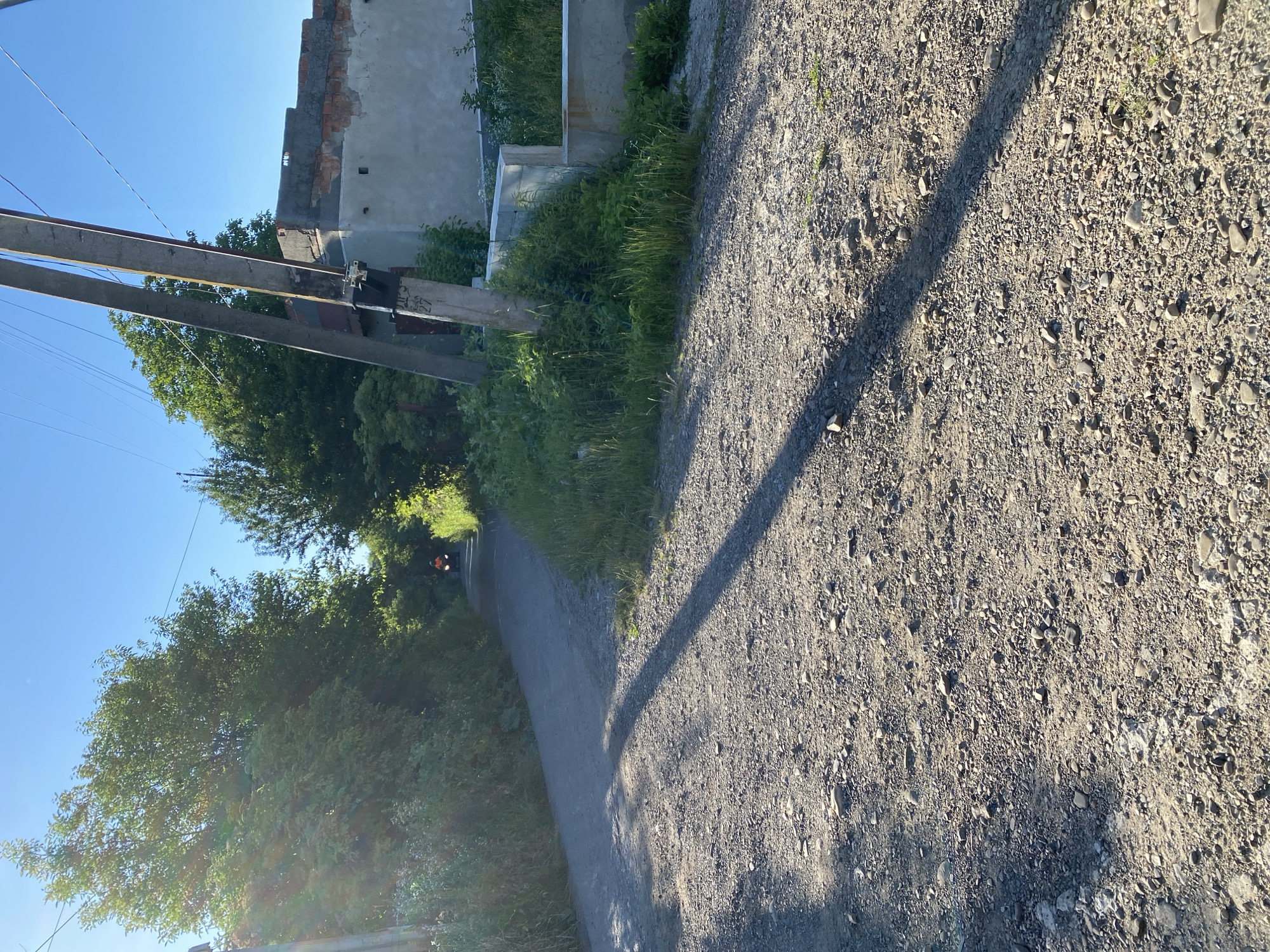 This is a photograph taken outdoors during the daytime, likely in the late afternoon given the long shadows. The scene depicts a dirt road bordered by lush green vegetation on the left and a gravelly, uneven surface on the right. In the distance, a person on a motorcycle or scooter, wearing orange and black, is visible on the road, suggesting movement or travel.
To the left of the road, there are trees and foliage, with a partial view of a building in the background. The building appears to be under construction or in disrepair, with exposed brickwork and concrete. Electrical poles with wires run along the left side of the frame, indicating the presence of infrastructure.
The lighting suggests a sunny day, casting distinct shadows across the gravel. The overall impression is one of a rural or semi-rural setting, possibly on the outskirts of a town or village. There are no discernible emotions or explicit textual elements in the image. The focus is on the landscape, the road, and the distant figure.