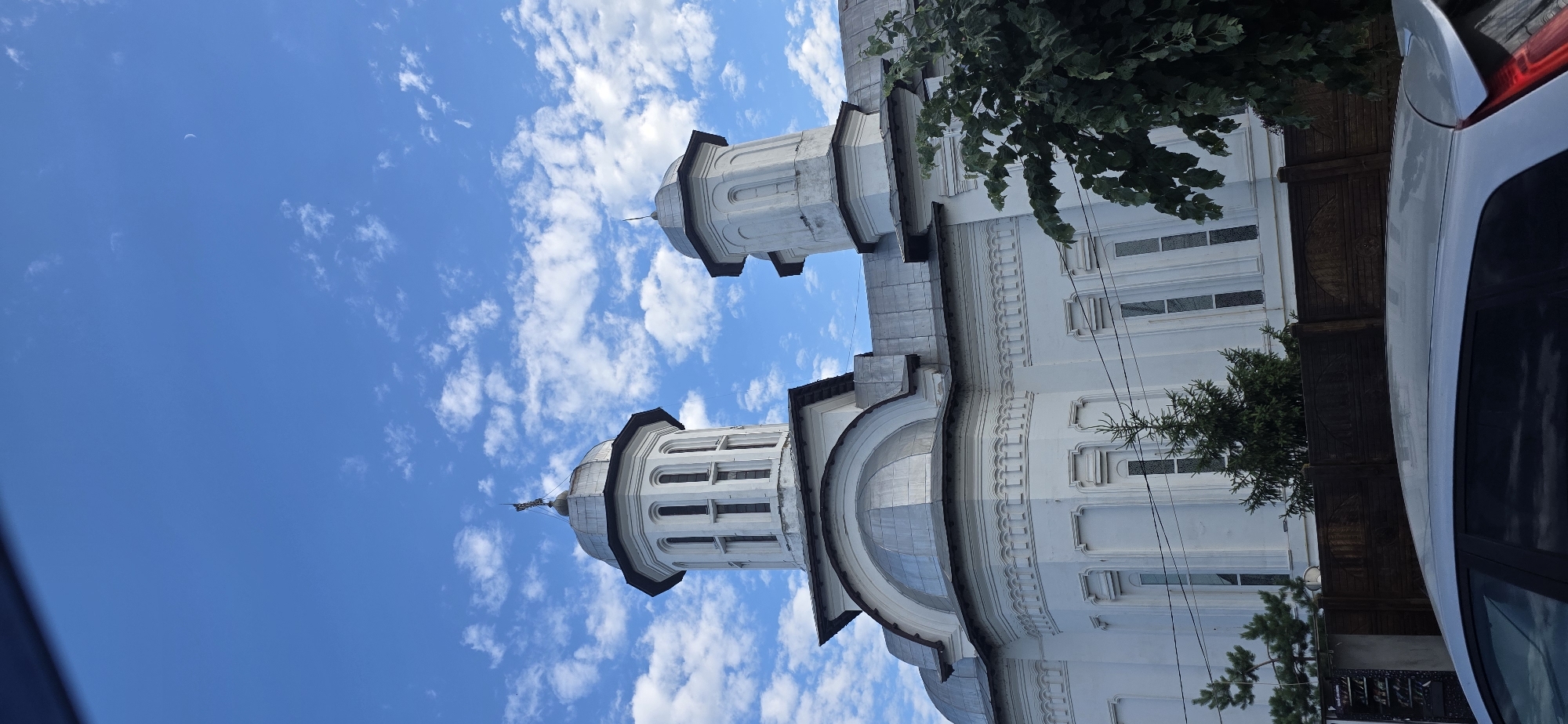 This is a vertically oriented, wide-angle, eye-level shot of a white church with two prominent bell towers under a bright blue sky filled with scattered white clouds. The church is a substantial building with ornate architectural details, including arched windows and decorative moldings along its facade. The domes of the bell towers are capped with dark material, and one appears to have a cross at its apex.
To the right of the church, a portion of a white car is visible, obscuring some of the building. Dense green foliage from trees overhangs the top right of the frame, partially obscuring the sky and the church. The scene appears to be set in a residential or urban area, as suggested by the presence of the car and some faint electrical wires. The bright sky and clear clouds indicate it is daytime, likely mid-morning or afternoon, with fair weather. There are no people visible in the frame. The image conveys a sense of stillness and grandeur associated with religious architecture. No visible text is present.