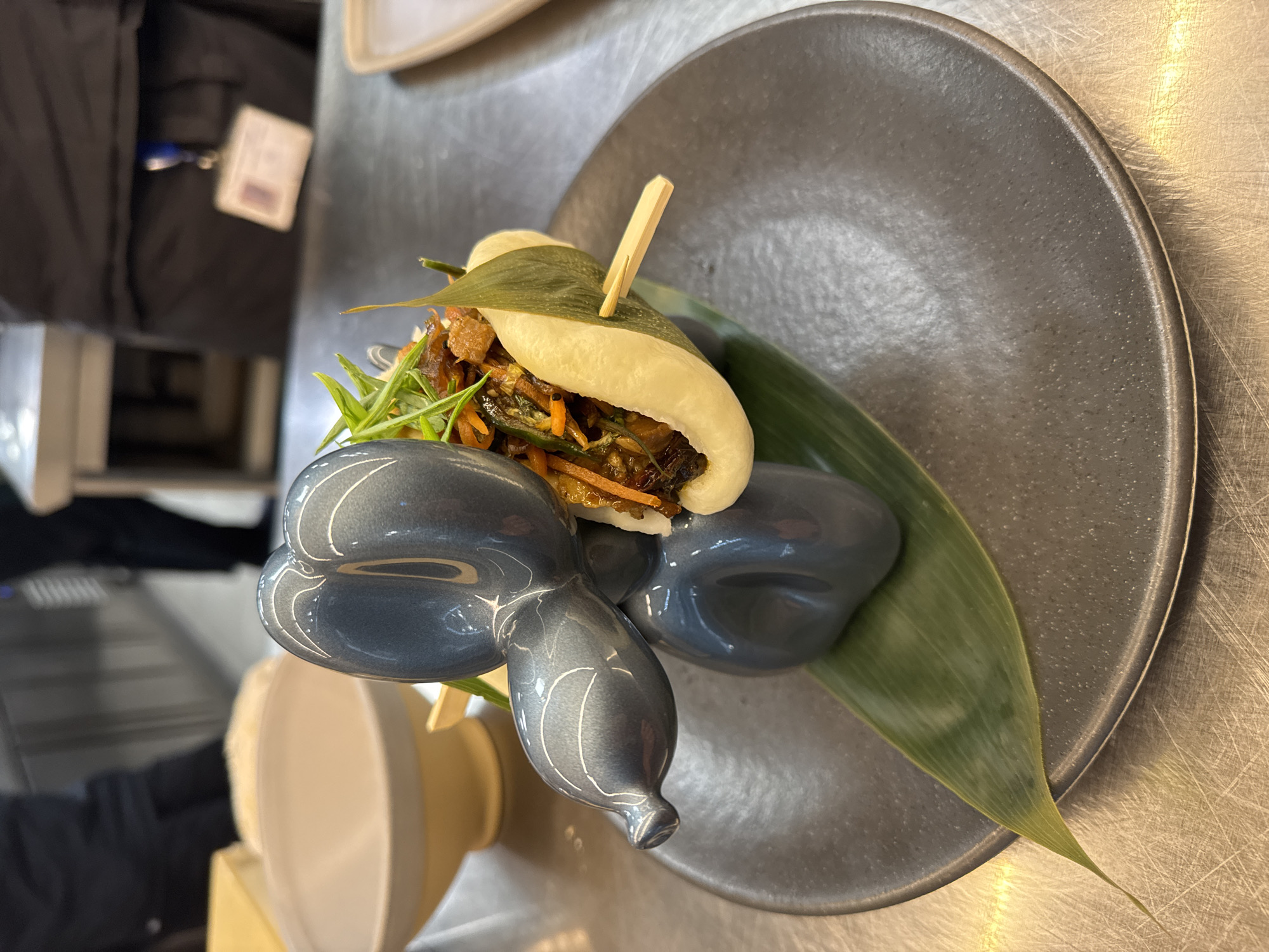 A close-up shot depicts a bao bun, skewered with a wooden pick, placed on a large green leaf, which rests on a dark grey, circular plate. Next to the plate is a grey balloon animal figurine. The plate sits on a metal surface with visible scratches. A blurry figure is partially visible on the left side of the frame, wearing a dark jacket with an ID tag. The image's origin or location in Sofia, Bulgaria, cannot be determined from the provided information.