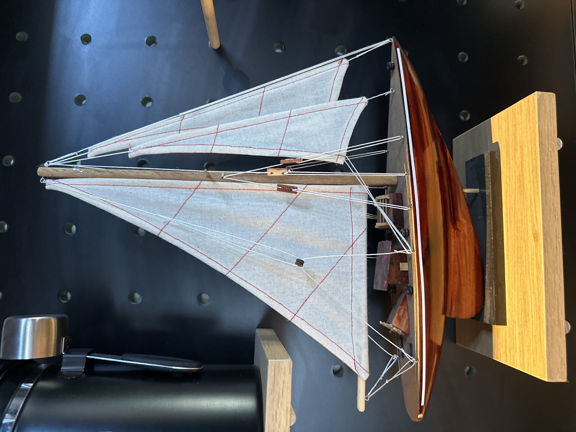 A detailed, forensic, matter-of-fact description of the scene:
The image depicts a model sailboat displayed on a light-colored wooden shelf. The sailboat has a wooden hull with a glossy, reddish-brown finish. It features two white sails with red stitching, supported by wooden masts and rigging. Small wooden structures are visible on the deck of the boat. The sailboat is positioned on a black base, which is in turn placed on the wooden shelf. To the left of the sailboat, a black cylindrical object with a silver metallic attachment is partially visible, also resting on a light-colored wooden shelf. The background is a dark, textured surface with a pattern of circular indentations. The lighting suggests an indoor environment, casting shadows on the background. There are no people or actions depicted. The scene is static and focuses on the display of the model sailboat. This image was taken in Da Nang, Vietnam.