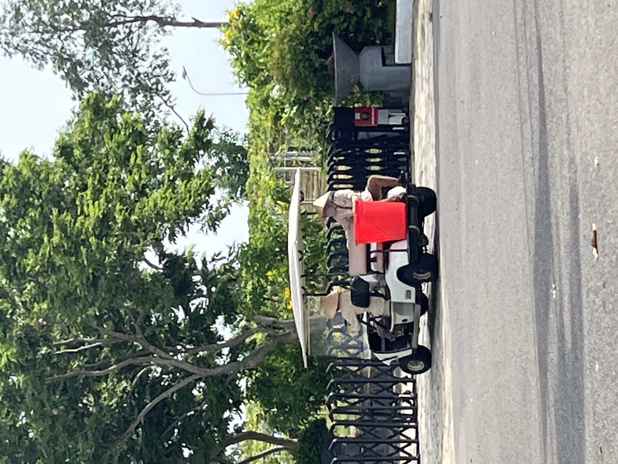 A white golf cart is parked on an asphalt road in front of a black metal gate. The golf cart is occupied by two people wearing conical hats. The person in the driver's seat is wearing a light-colored shirt and brown pants. The person in the passenger seat is wearing a light-colored shirt and appears to be looking down. A large red plastic container is visible in the back of the golf cart. Behind the gate, there are lush green trees and bushes. A street lamp is visible in the background. The scene appears to be a sunny day. The location is Phu Thuy Ward, Vietnam.
