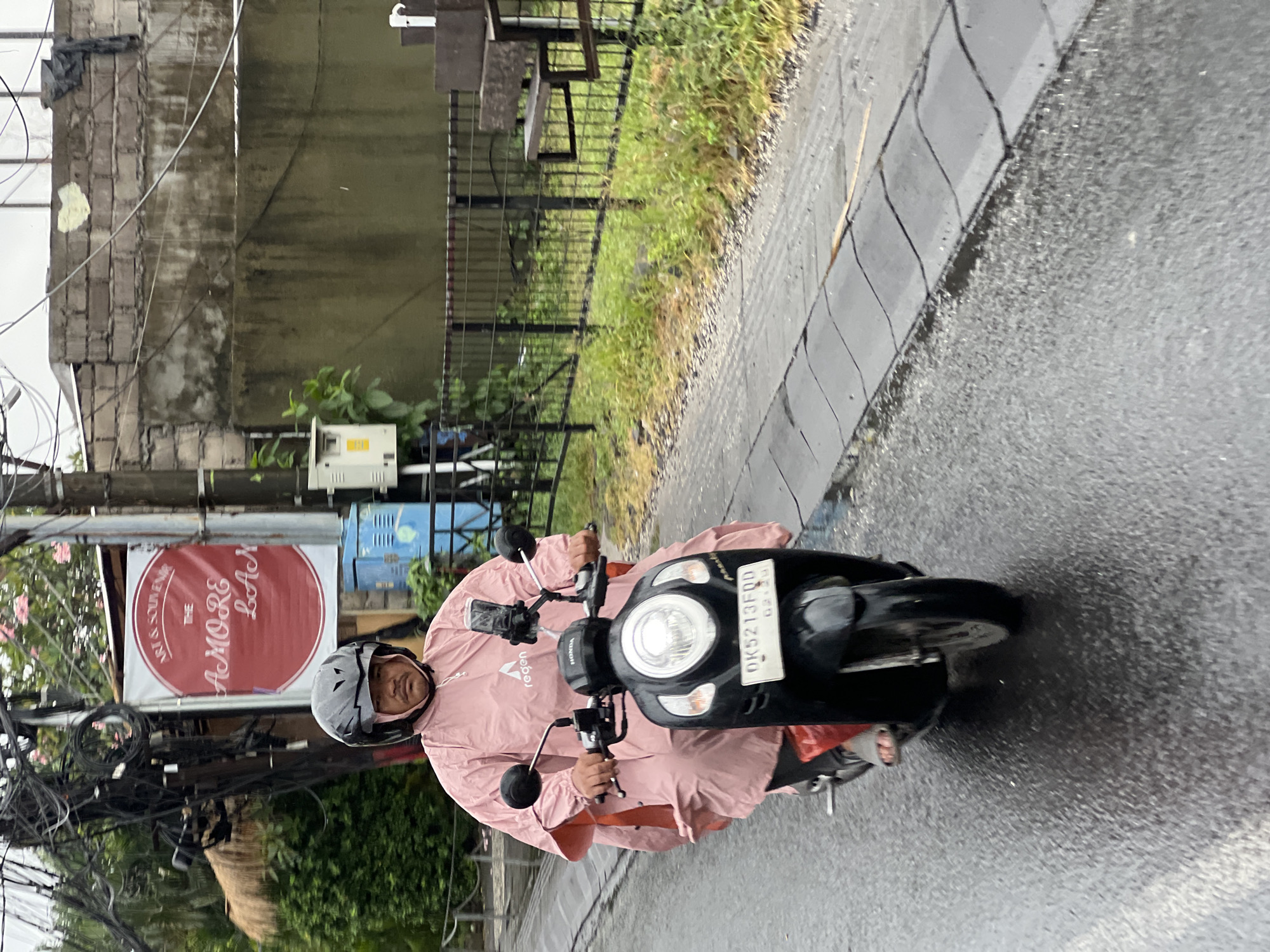 In North Kuta, Indonesia, a person wearing a pink poncho and a helmet rides a black scooter on a wet, paved road. The scooter has a license plate that reads "DK5213FQD". A cell phone is mounted on the handlebars. Behind the rider, a sign reads "ART & SOUVENIRS THE AMORE LAN". Electrical wires hang overhead, and a concrete wall with some staining is visible in the background. Green vegetation and a metal fence are also present. The ground is wet, indicating recent rain.