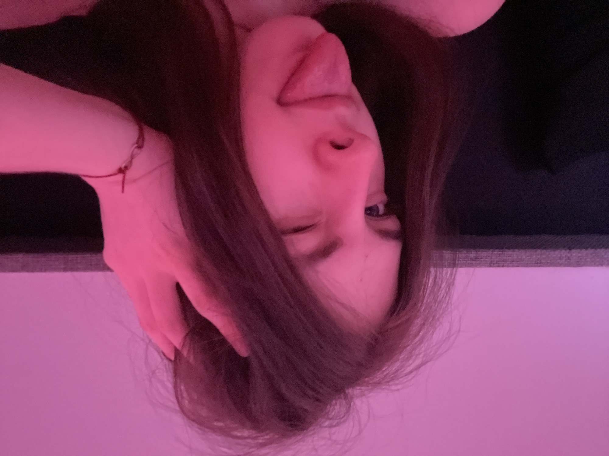A person with long brown hair is sticking their tongue out and winking in an upside-down close-up image, bathed in pink light. One hand is near their head, possibly touching the hair. The location is not discernible from the image.