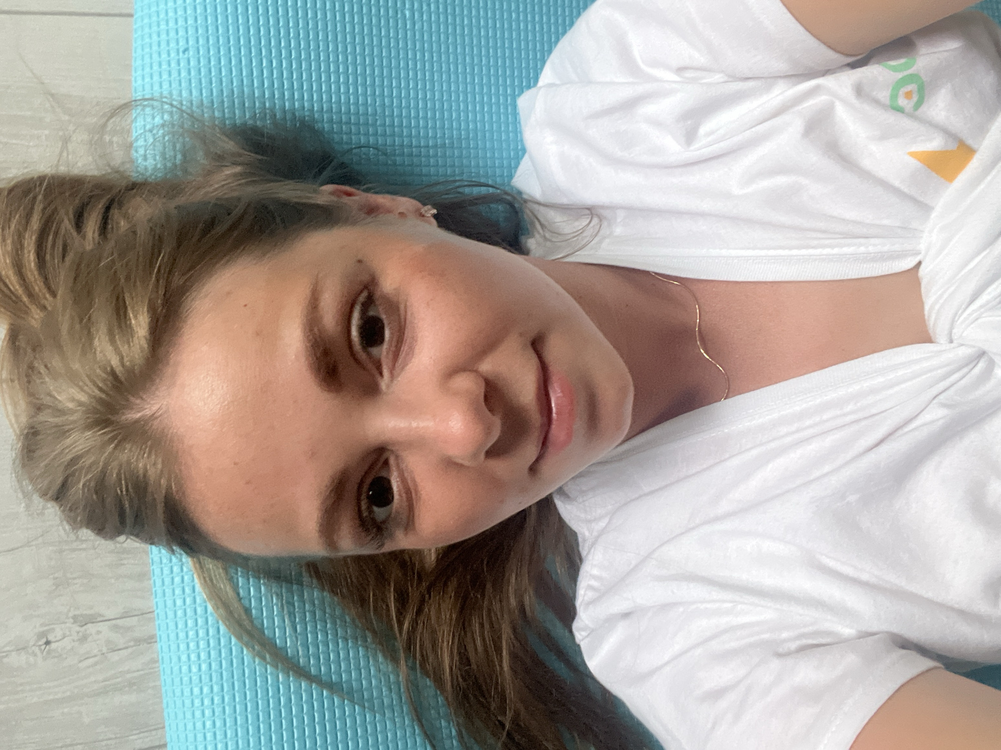 A young woman lies on her back on a blue yoga mat, which is placed on a light gray, wooden-patterned floor. She is wearing a white t-shirt with a green and yellow graphic on the upper chest. A thin gold chain necklace is visible around her neck. Her blonde hair is spread out around her head. The image is a close-up, focusing on her face and upper body. She is looking at the camera with a slight smile. The lighting suggests it might be daytime, possibly indoors with natural light. The overall mood seems relaxed and casual. There is no visible text in the image apart from the graphic on her t-shirt, which is indistinct.