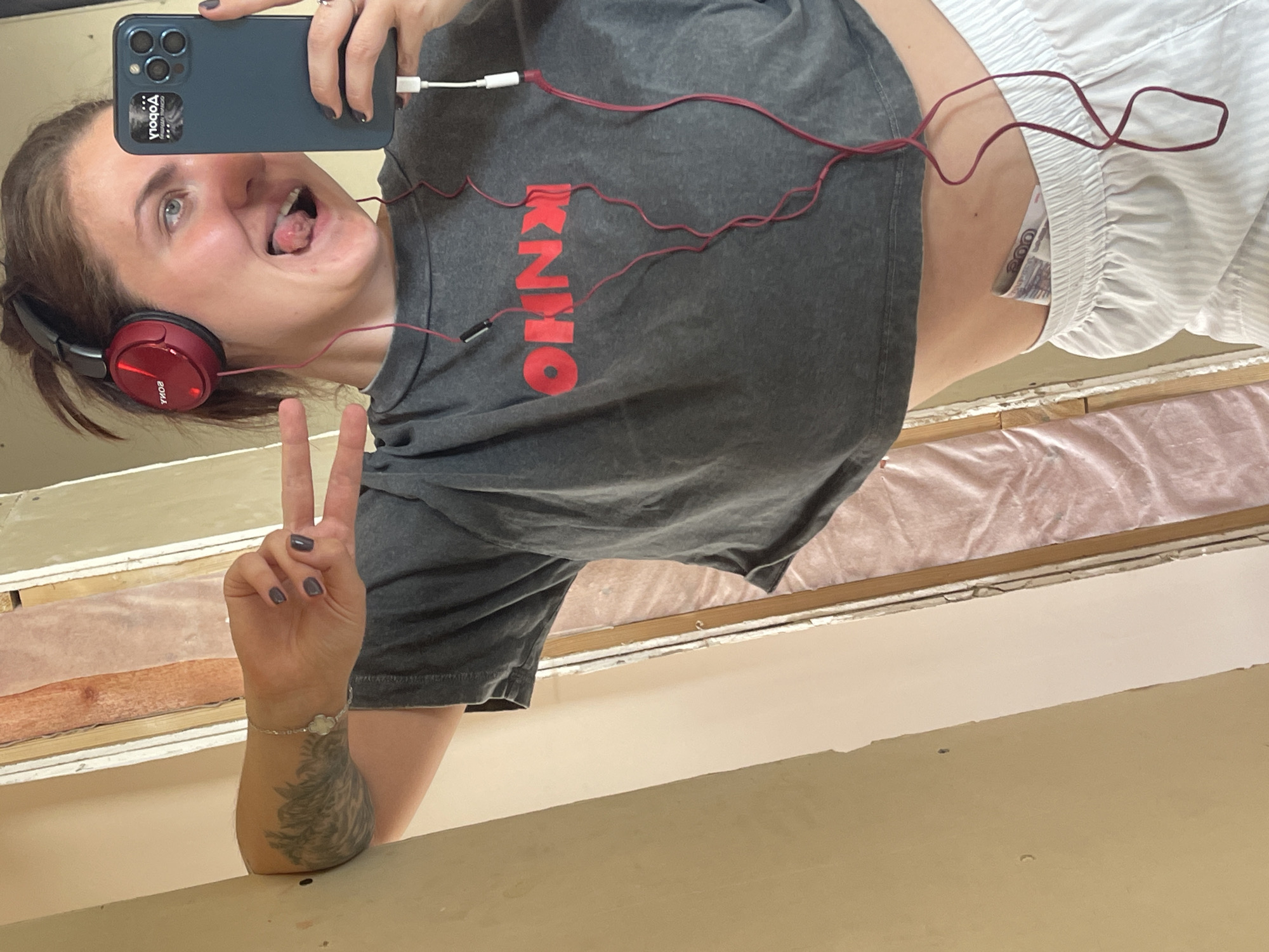 A young woman is taking a selfie. She is wearing red headphones, a grey crop top with red lettering, and white striped shorts. She is holding a blue smartphone with a triple-lens camera, and her tongue is sticking out. She is making a peace sign with her left hand, which has a wrist tattoo. A pair of red wired headphones are connected to the phone. A bill is visible in her shorts pocket. The setting appears to be indoors, possibly a room under renovation, with exposed wood and insulation. This image cannot be used to reliably determine any location in the city of Мосметаллоконструкция Онт, Russia.